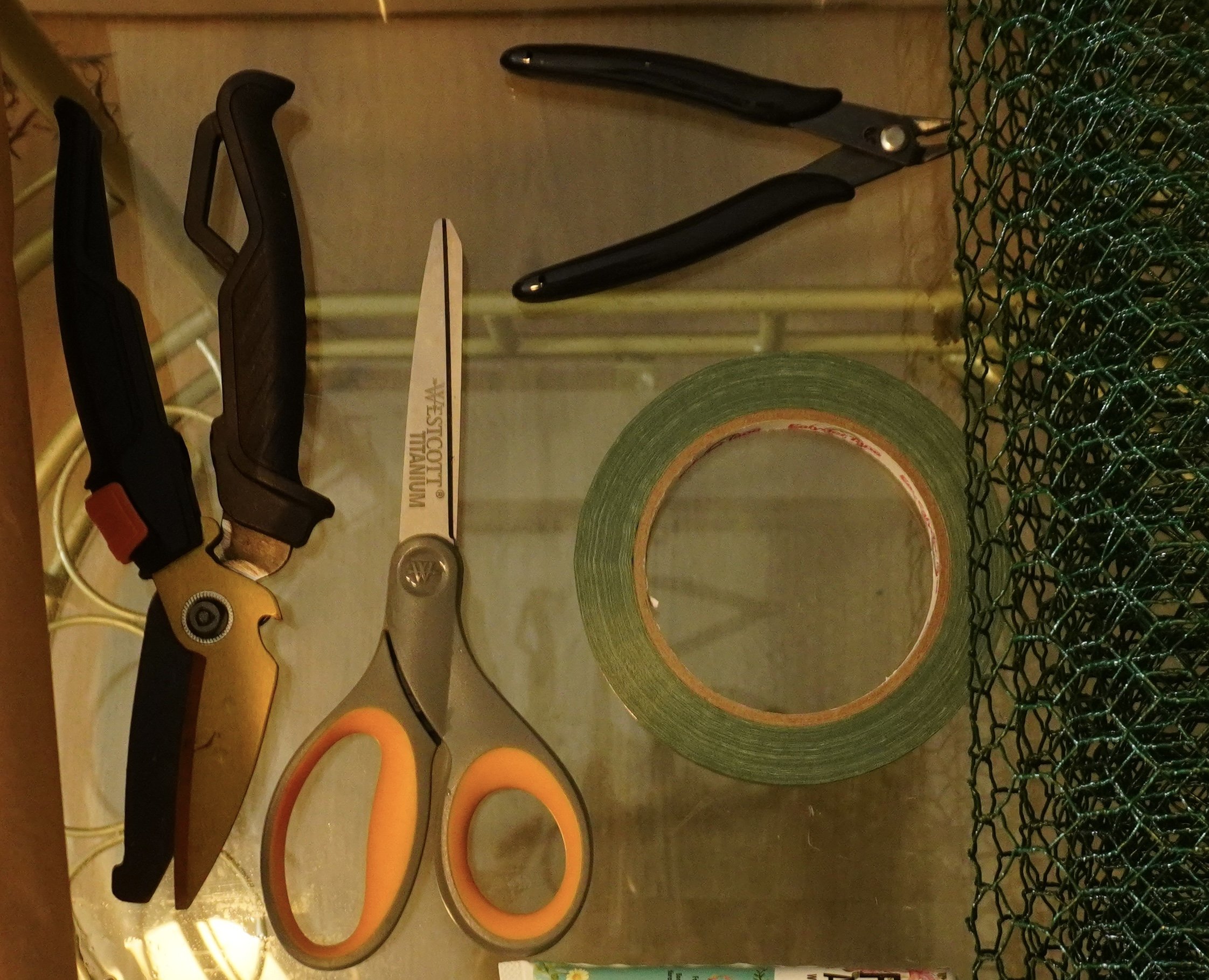Tools and supplies on a kitchen countertop, including a handsaw, a miniature saw, a roll of green painter's tape, orange rubber gaskets, and a piece of green wire mesh.