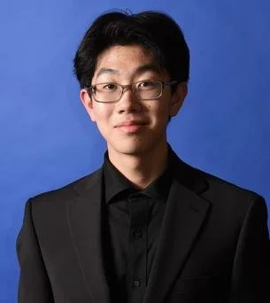 Young man with glasses wearing a black suit and shirt, posing against a blue background.