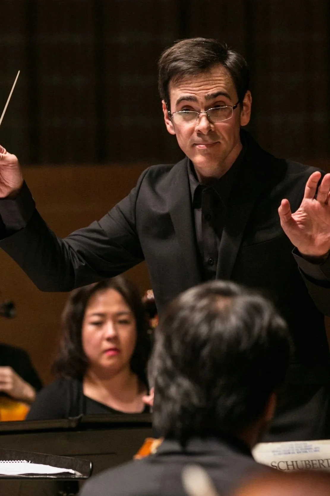Man conducting orchestra, wearing glasses and black suit, with a serious expression, behind a woman with dark hair, in a concert hall.