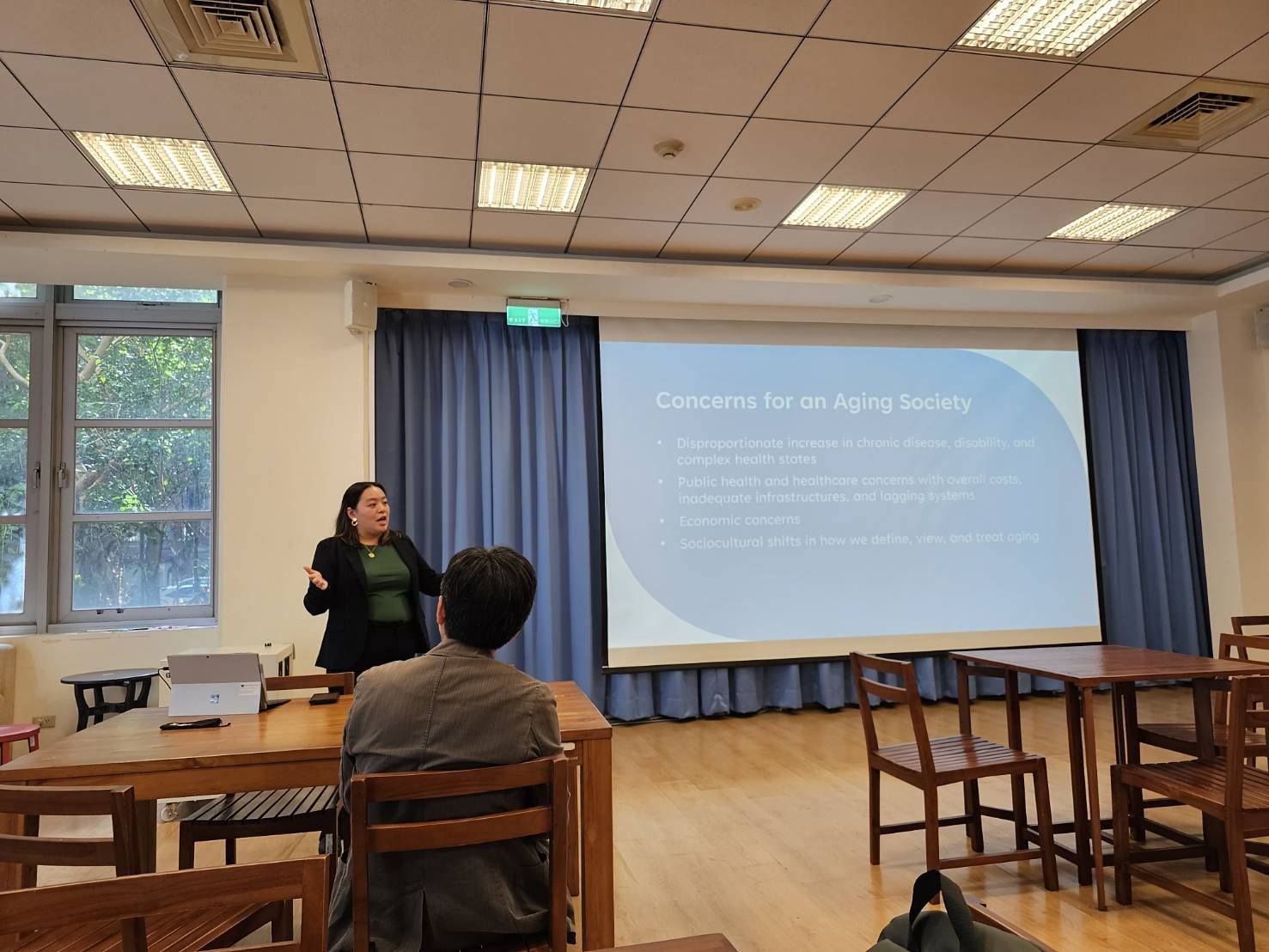 Dr. Cassie Lopez-Jeng giving a lecture. She's standing at the front of the room next to a slide titled Concerns for an Aging Society.