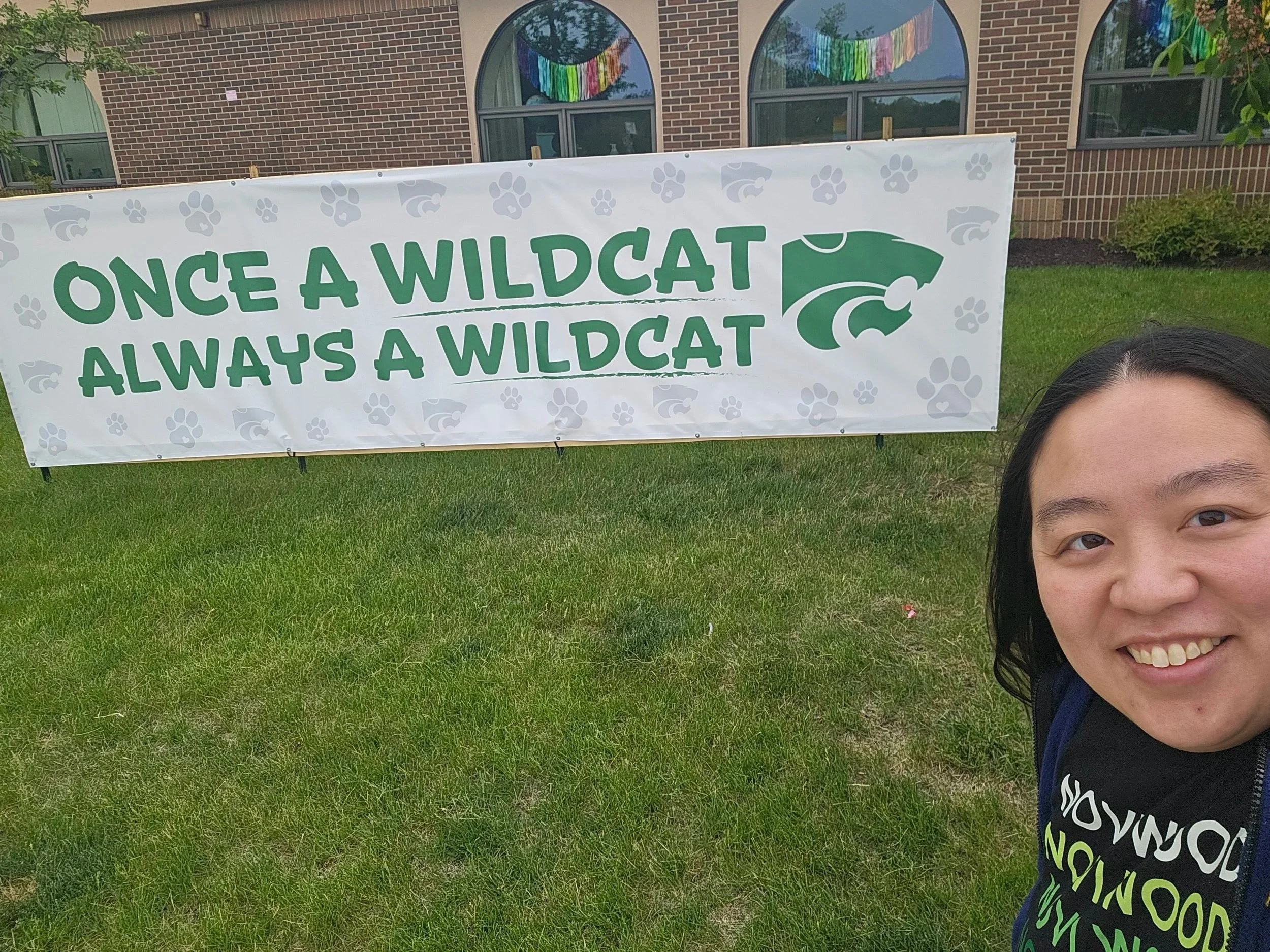 Dr. Cassie Lopez-Jeng stands next to a sign in front of Novi Woods that says "Once a Wildcat, Always a Wildcat"