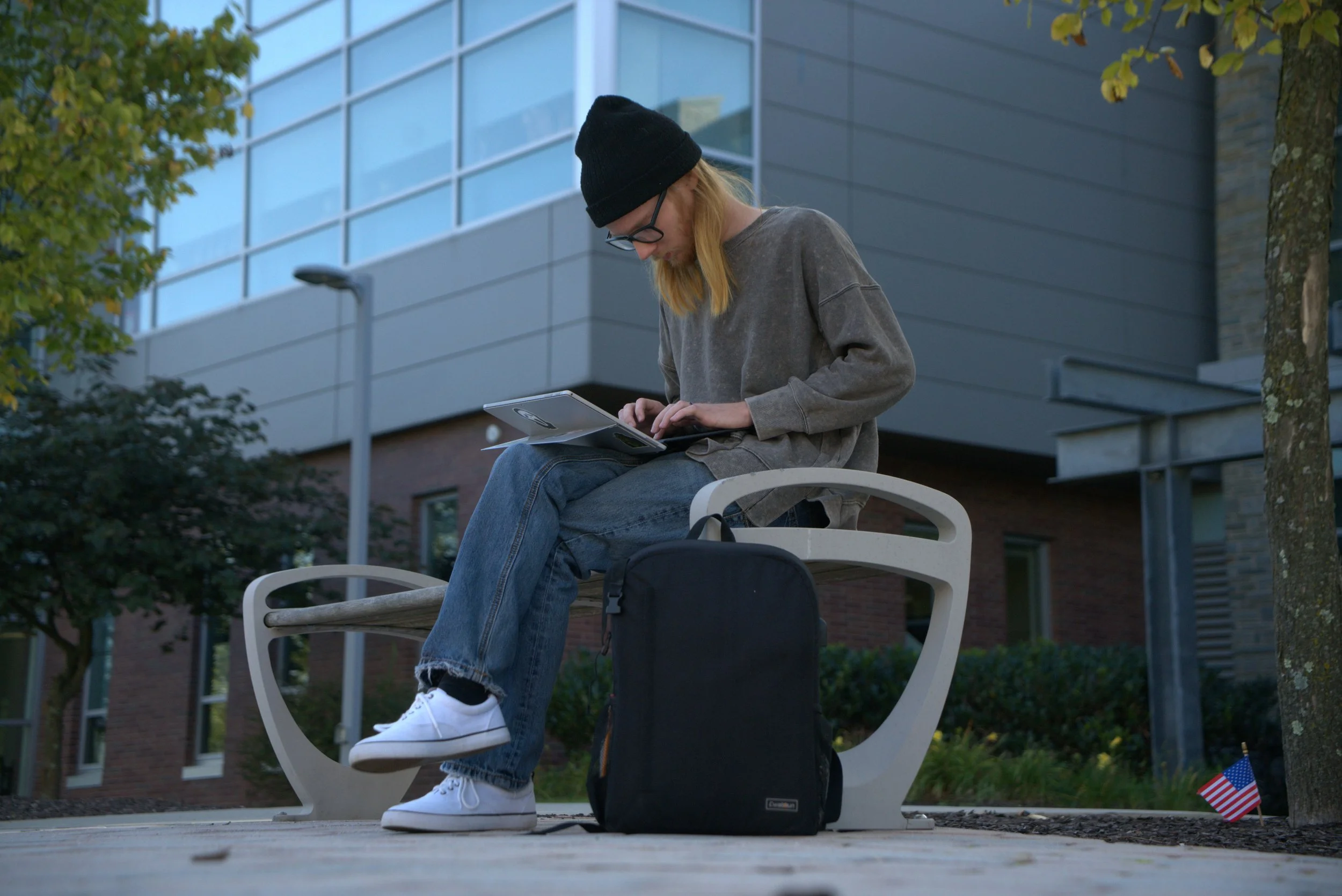A person with long blonde hair, black beanie, glasses, gray oversized sweatshirt, and jeans sitting on a white bench outdoors, looking at a tablet. They have a black backpack and an American flag on the ground nearby. There are trees and modern buildings in the background.