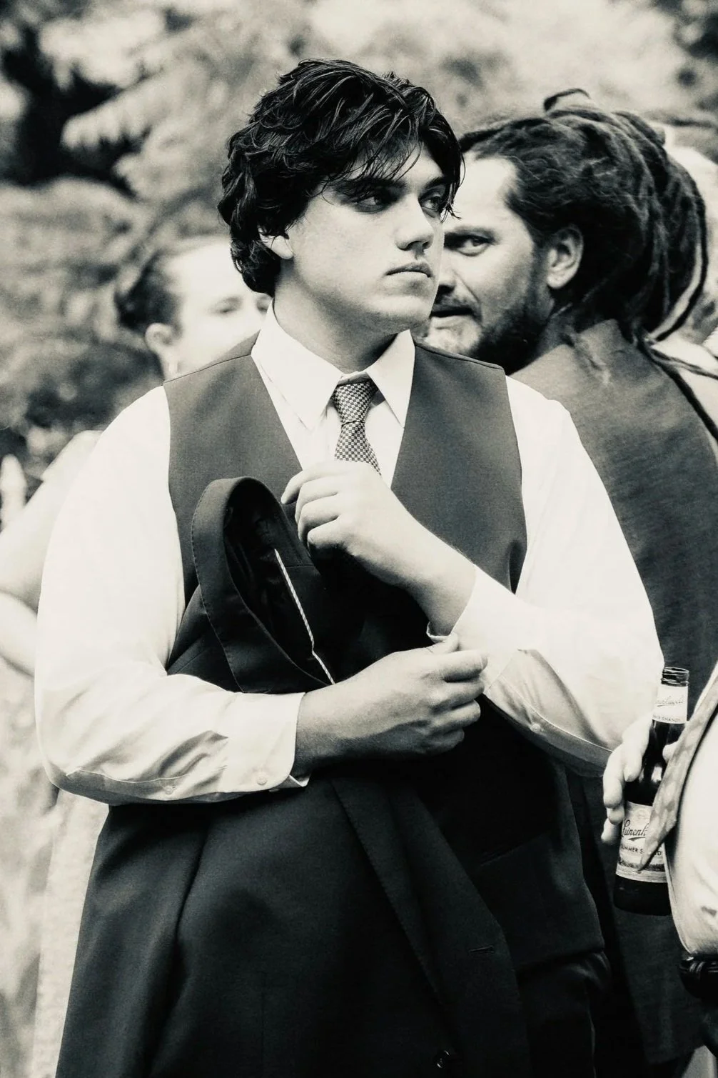 Young man with dark, hair styled in a vintage fashion, wearing a white shirt, vest, and tie, holding a jacket and standing outdoors surrounded by people.