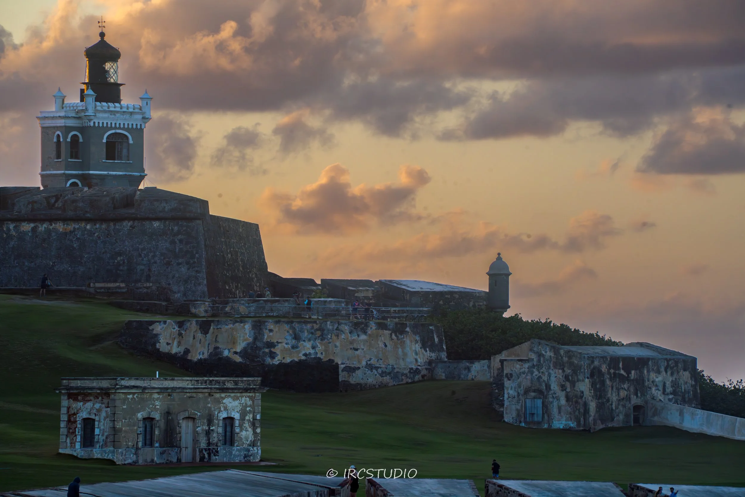 A historic fortress with a lighthouse tower overlooking the sea at sunset, with a cloudy sky and people walking along the walls.