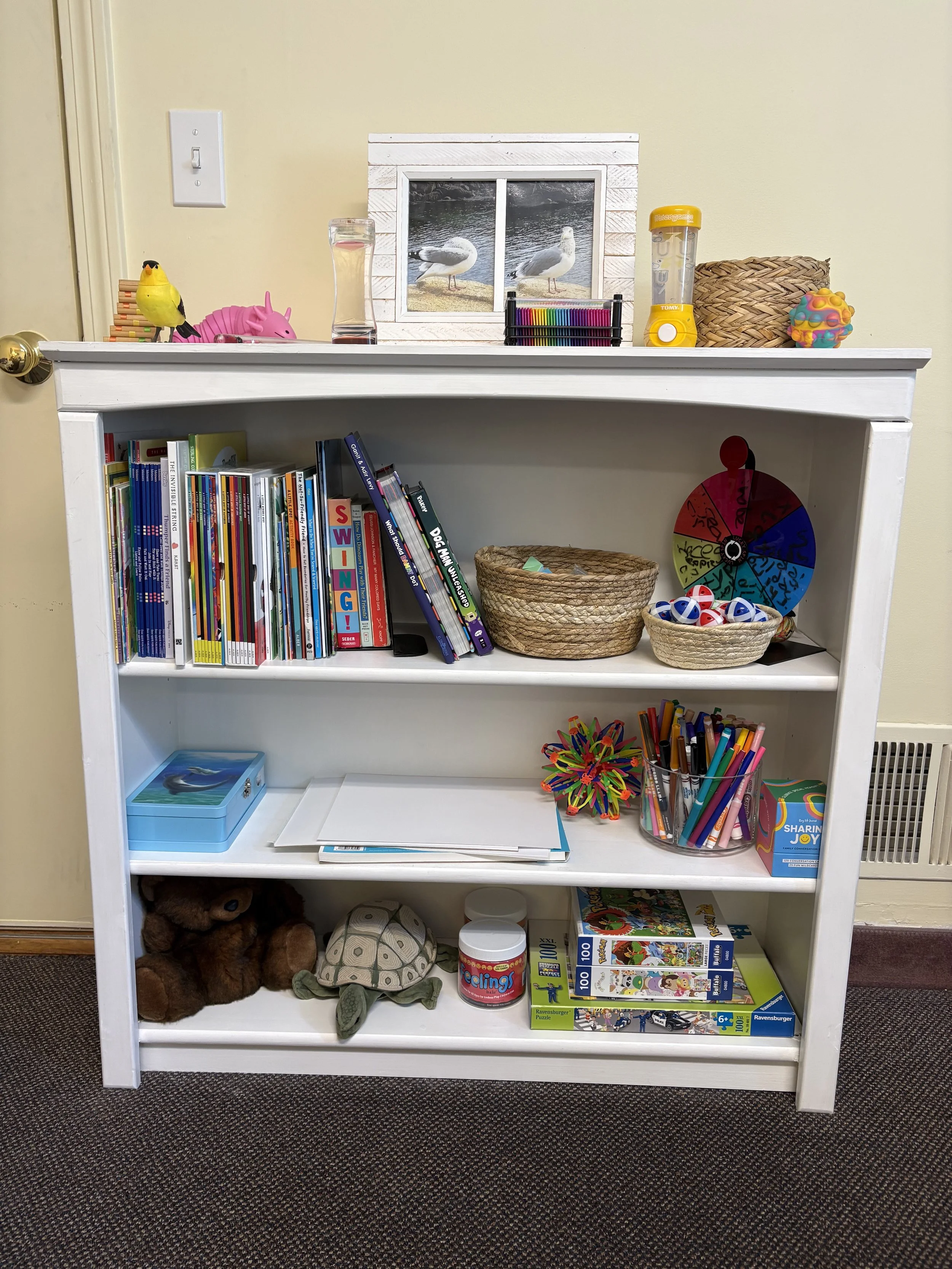 A white bookshelf with four shelves contains children's books, toys, puzzles, art supplies, and a stuffed animal. The top of the bookshelf has a small window with drawings of two ducks by a lake, and decorative items including a bird toy, a pink pig, a lamp, a colorful wheel, and a container of sensory putty.