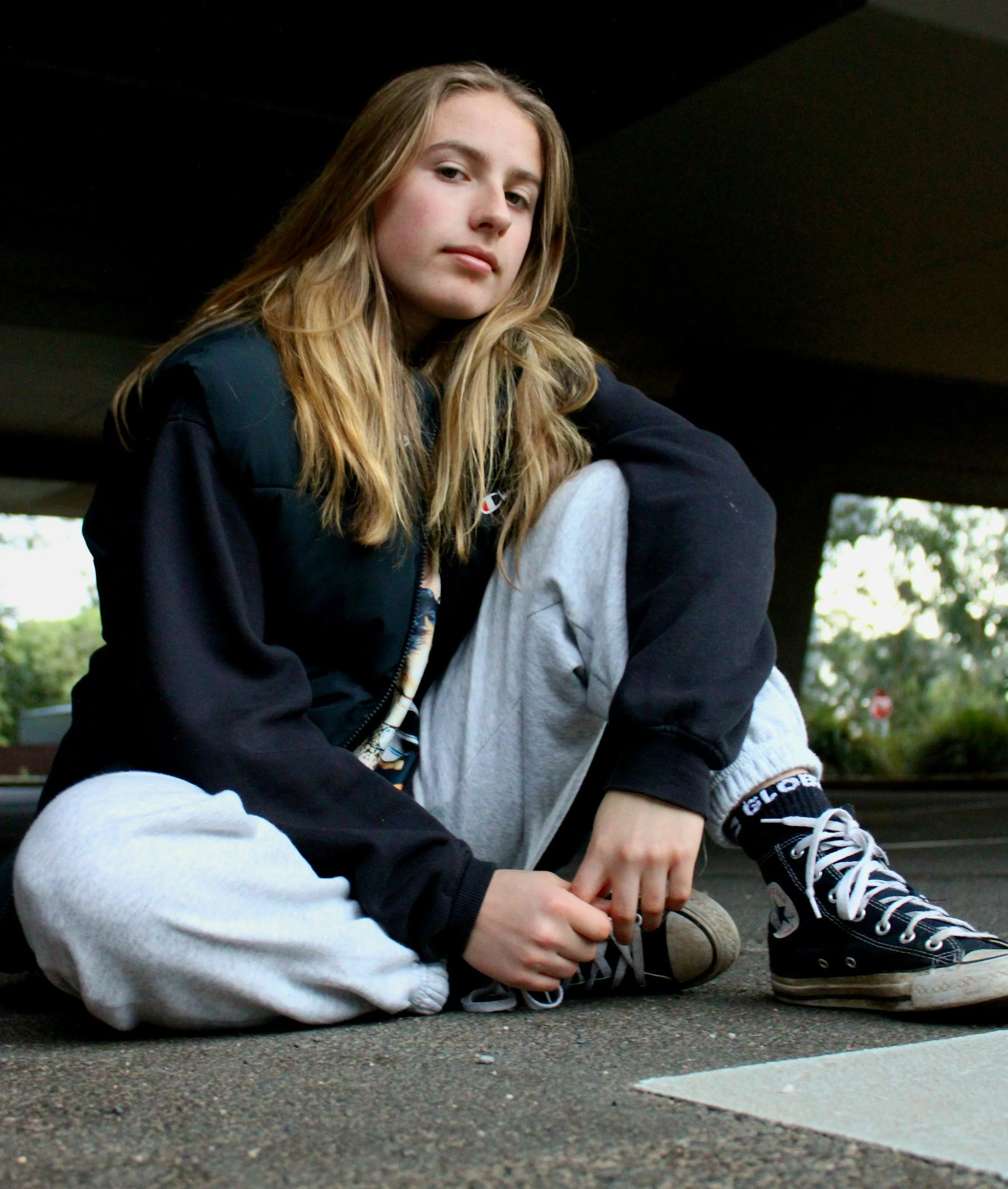 A teen with long blonde hair sitting on the ground in an outdoor space wearing sneakers, black sweatshirt and gray sweatpants.
