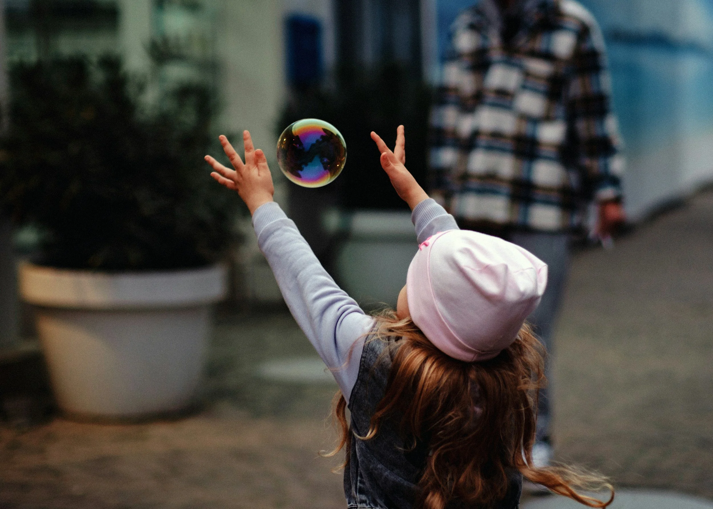 Child with long hair wearing a pink hat and dark vest, reaching up towards a colorful soap bubble in an urban outdoor setting.
