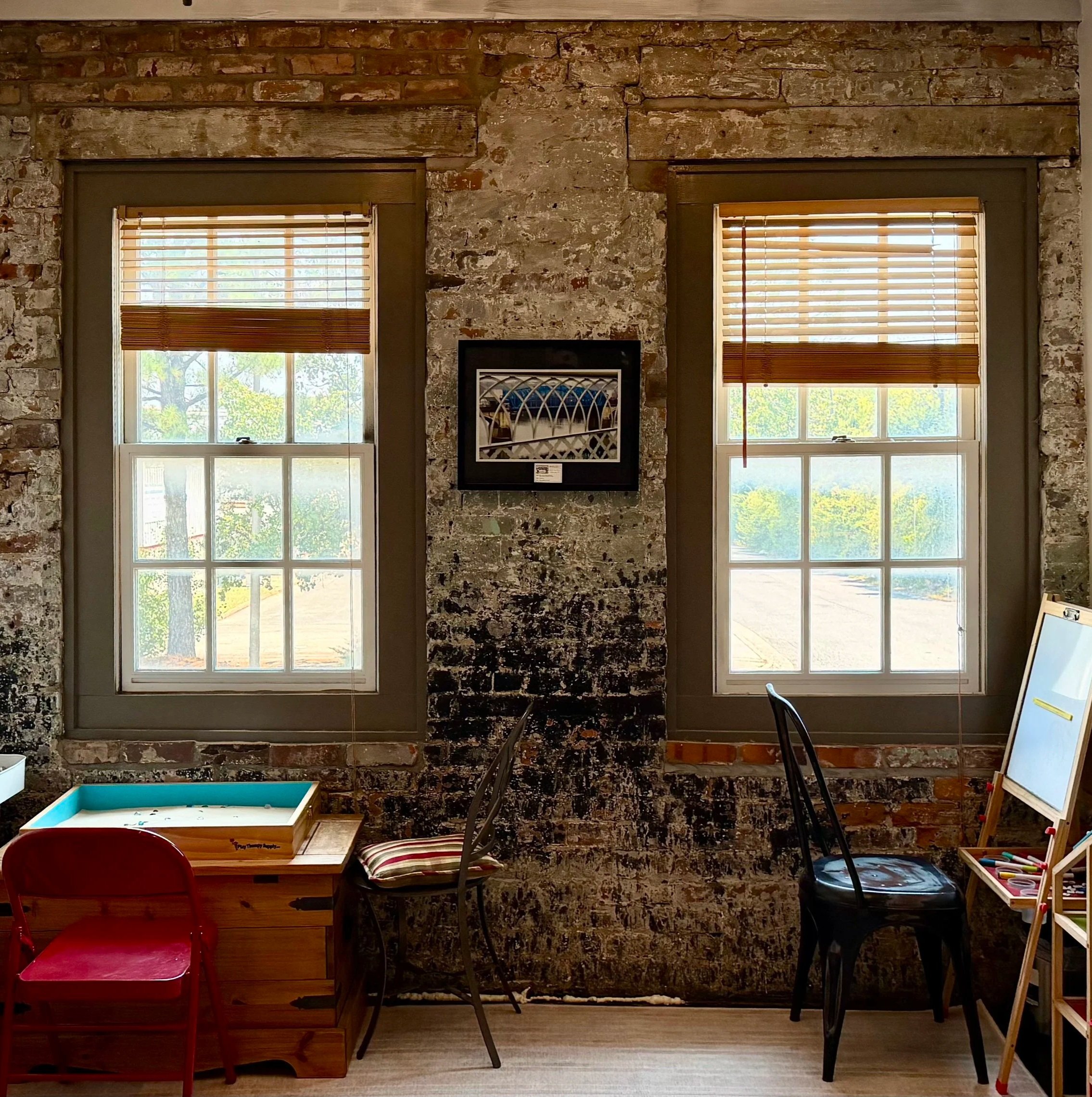 Rustic brick-walled room with two windows, wooden blinds, framed artwork, chairs, a sand tray therapy table, and a children's easel