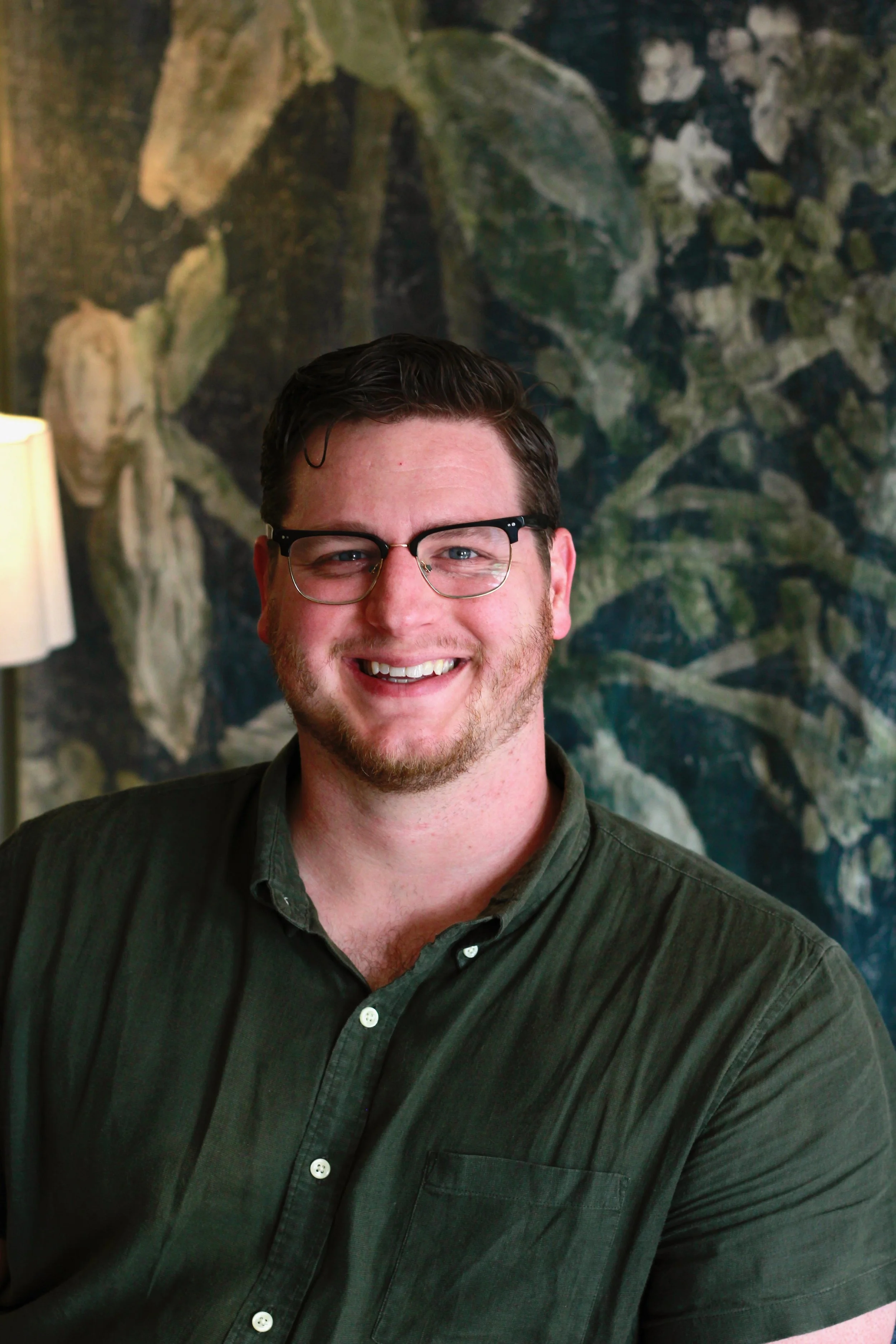 Andrew Parks, licensed counselor & Registered Play Therapist wearing glasses and a dark green shirt, sitting in his therapy chair in front of a forest mural.