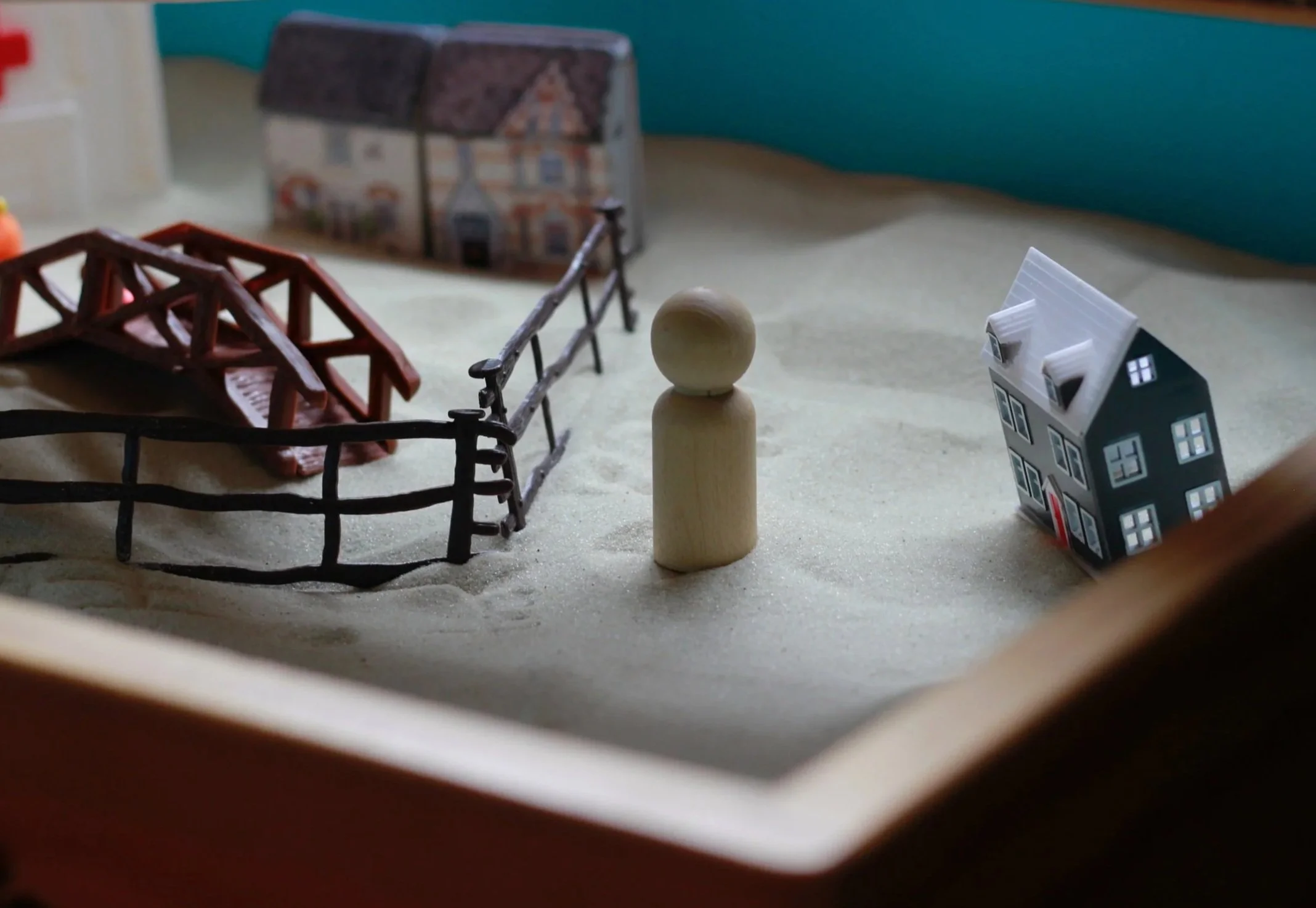 Miniature Sand Tray Therapy scene with a wooden peg person, model house, bridge, and black fence.