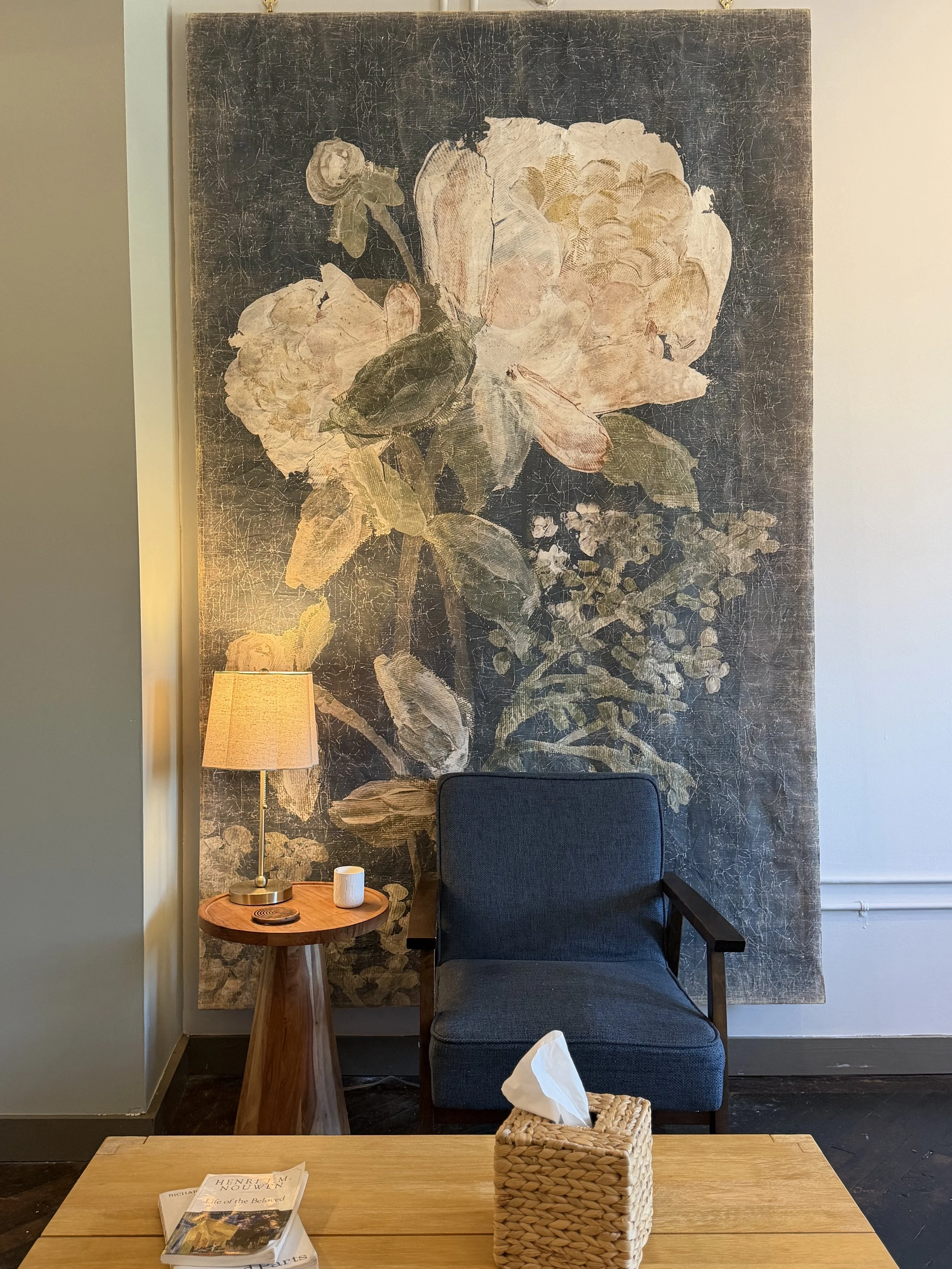 Cozy sitting area with a blue armchair, floral wall art, side table and lamp, and a woven tissue box on a wooden coffee table