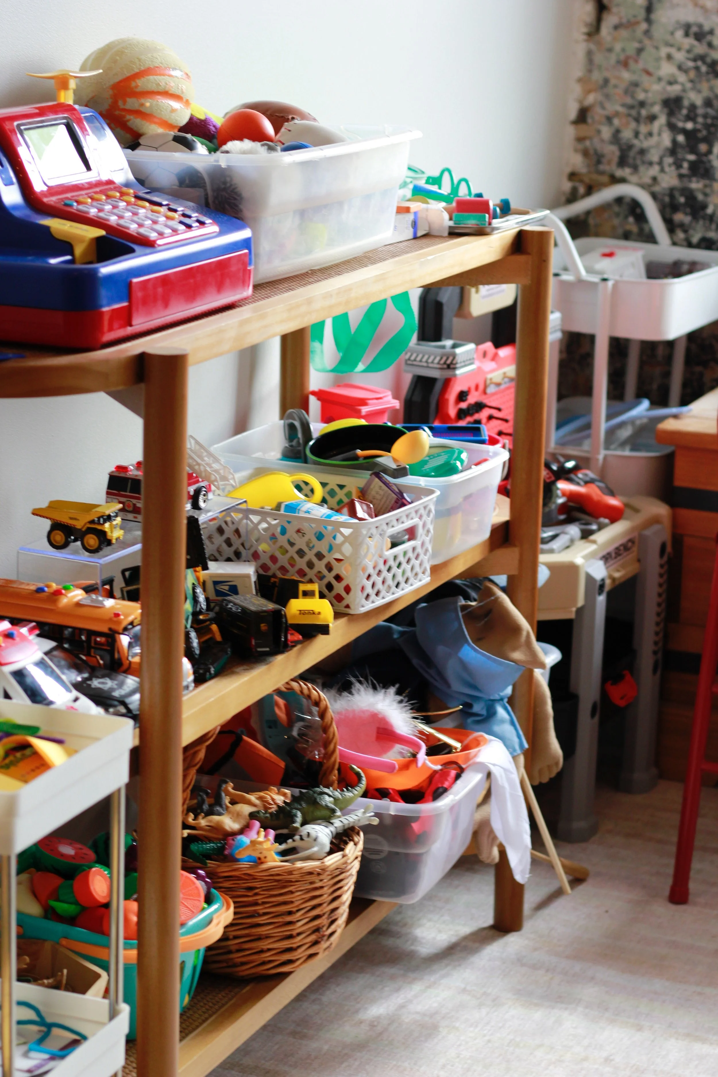Wooden shelving unit filled with organized bins and baskets of children's toys, including toy vehicles, sports balls, action figures, and play food -- all use to facilitate Play Therapy and helping children heal.