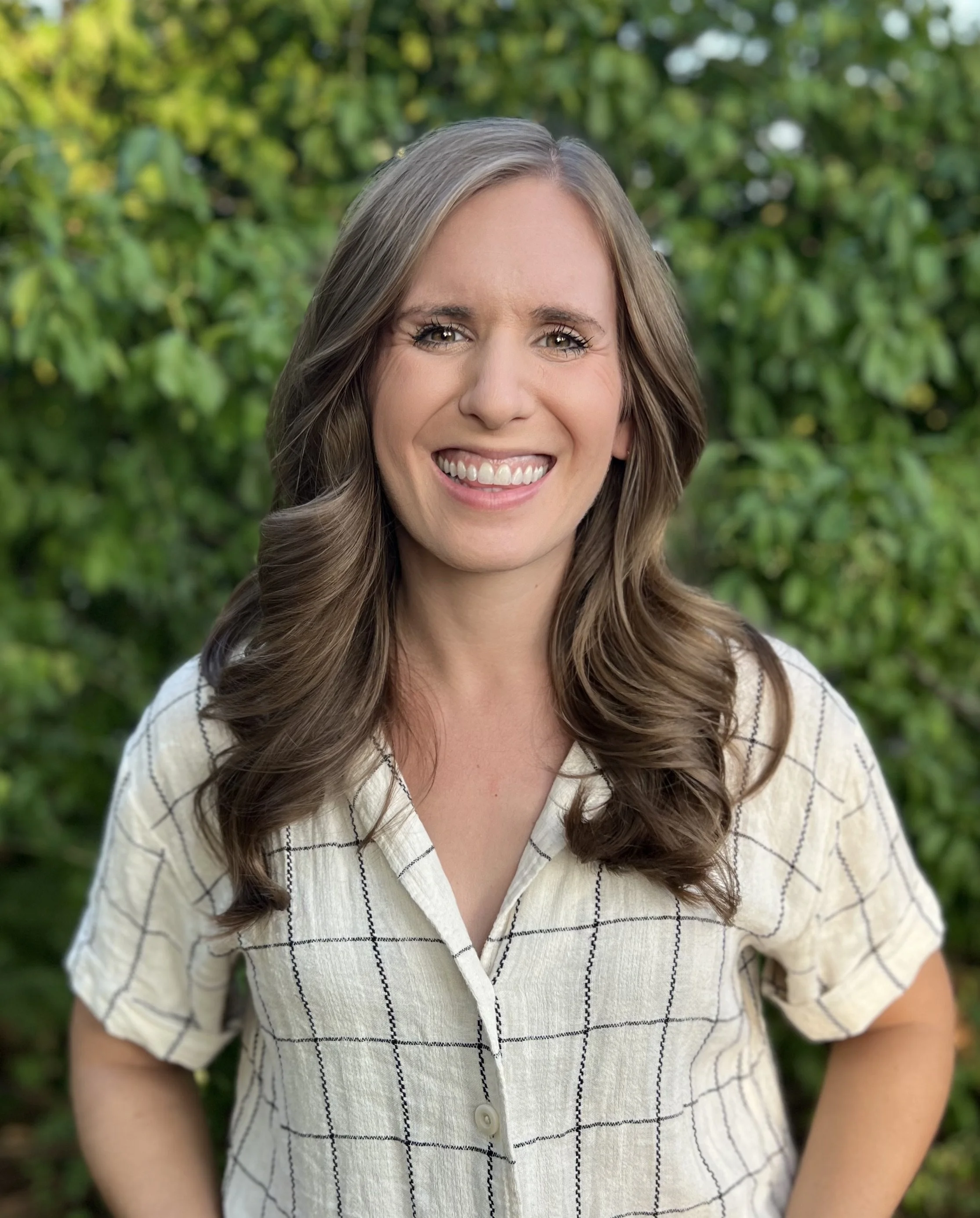 Smiling woman with long brown hair in a white checkered shirt outdoors against a leafy green backdrop. This is Mary Grace Parks, a counselor who specializes in warm, compassionate care for postpartum moms & those navigating perinatal mental health.