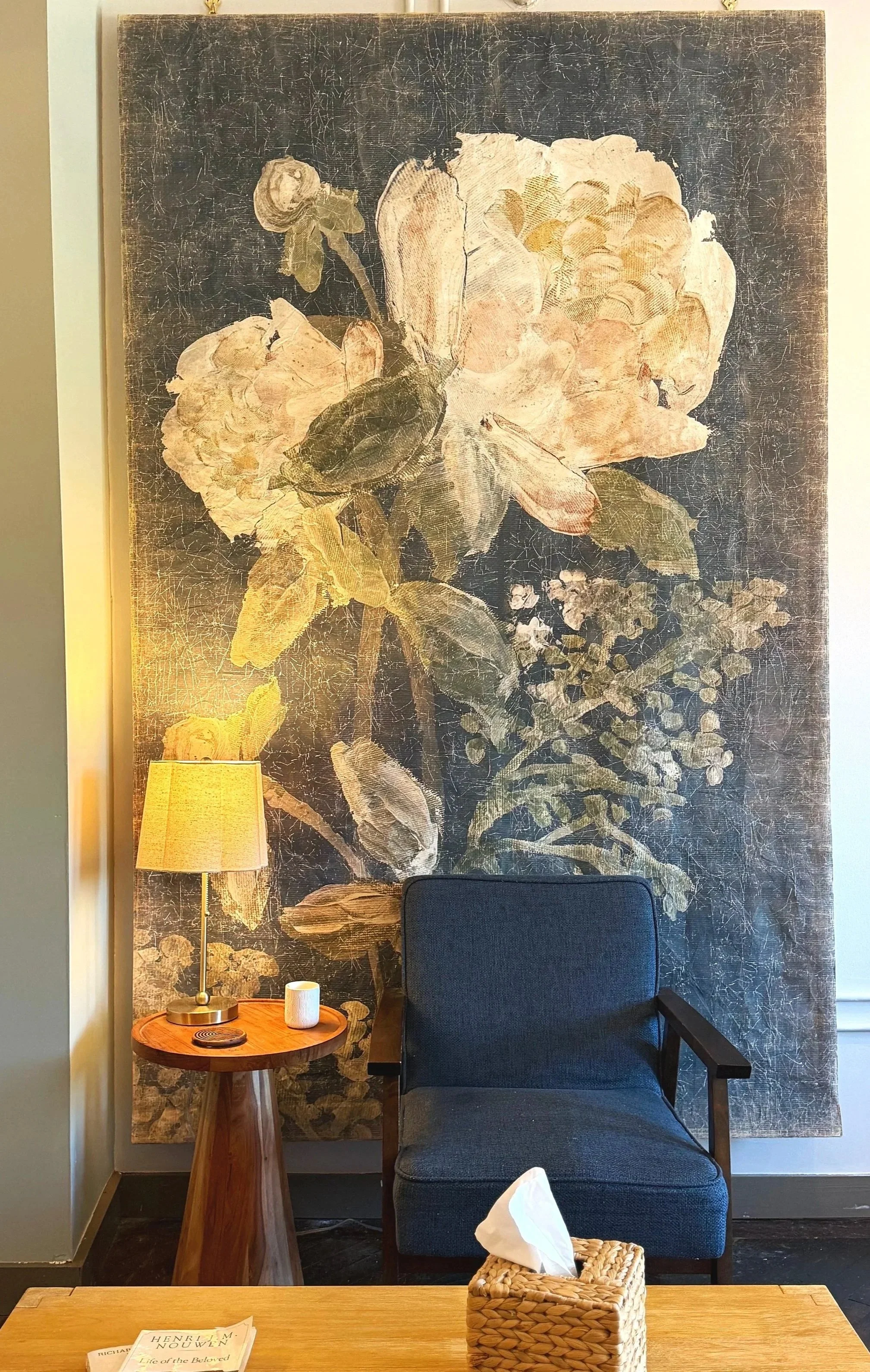 Cozy therapist's chair with a blue armchair, floral wall mural, floor lamp, round side table, books, and woven tissue basket.