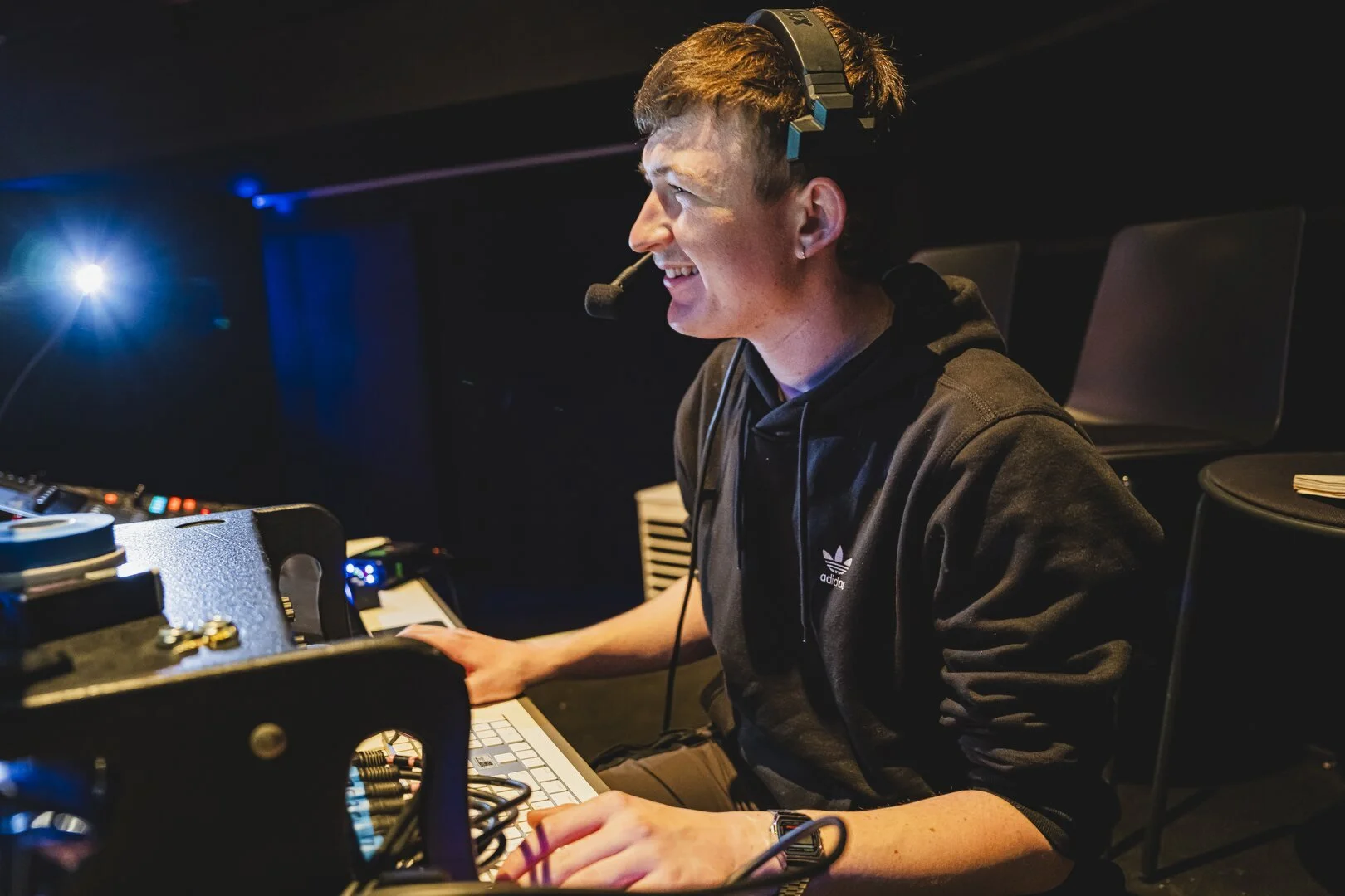 A young man with a headset and microphone sitting at a sound production console in a dark studio with blue lighting.