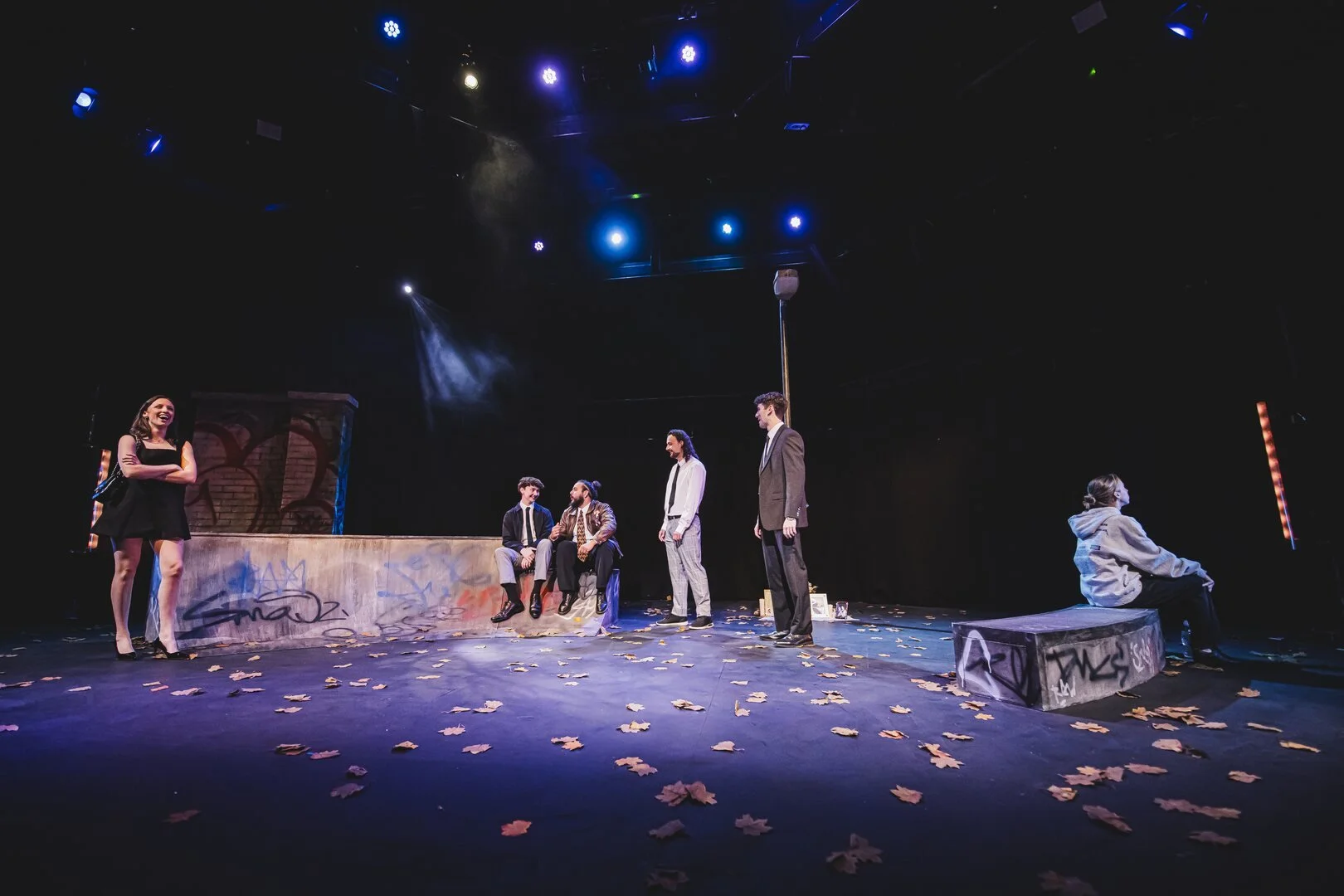 A stage play featuring six actors, four standing and two sitting, with graffiti-covered props and scattered leaves on the floor, under colorful stage lighting.