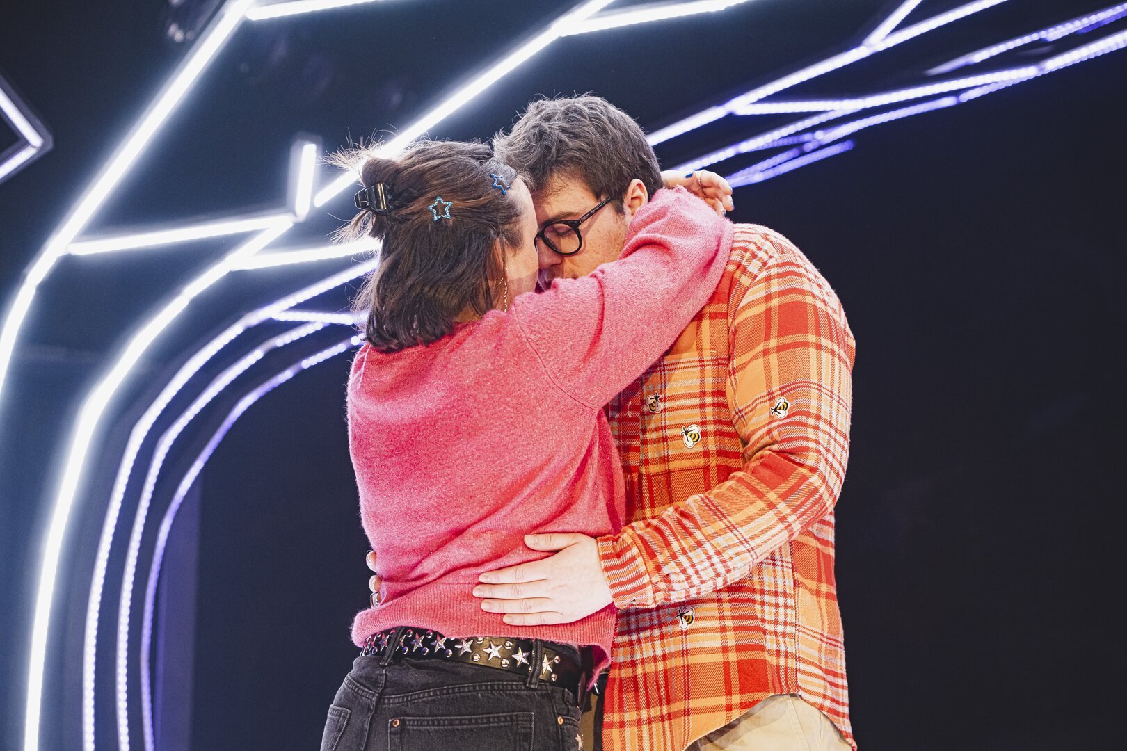 Two people hugging in front of a neon light tree, one wearing a pink sweater with star clips in hair, the other in a plaid jacket with bee pins.