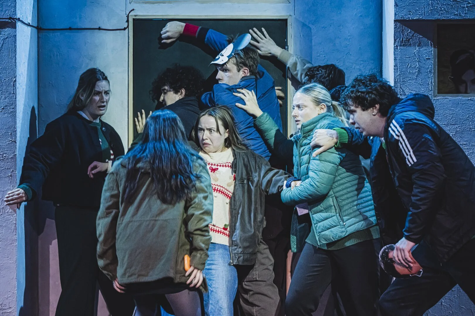 A group of people in a tense confrontation outside a door, some pushing and grabbing each other, with distressed and angry expressions.
