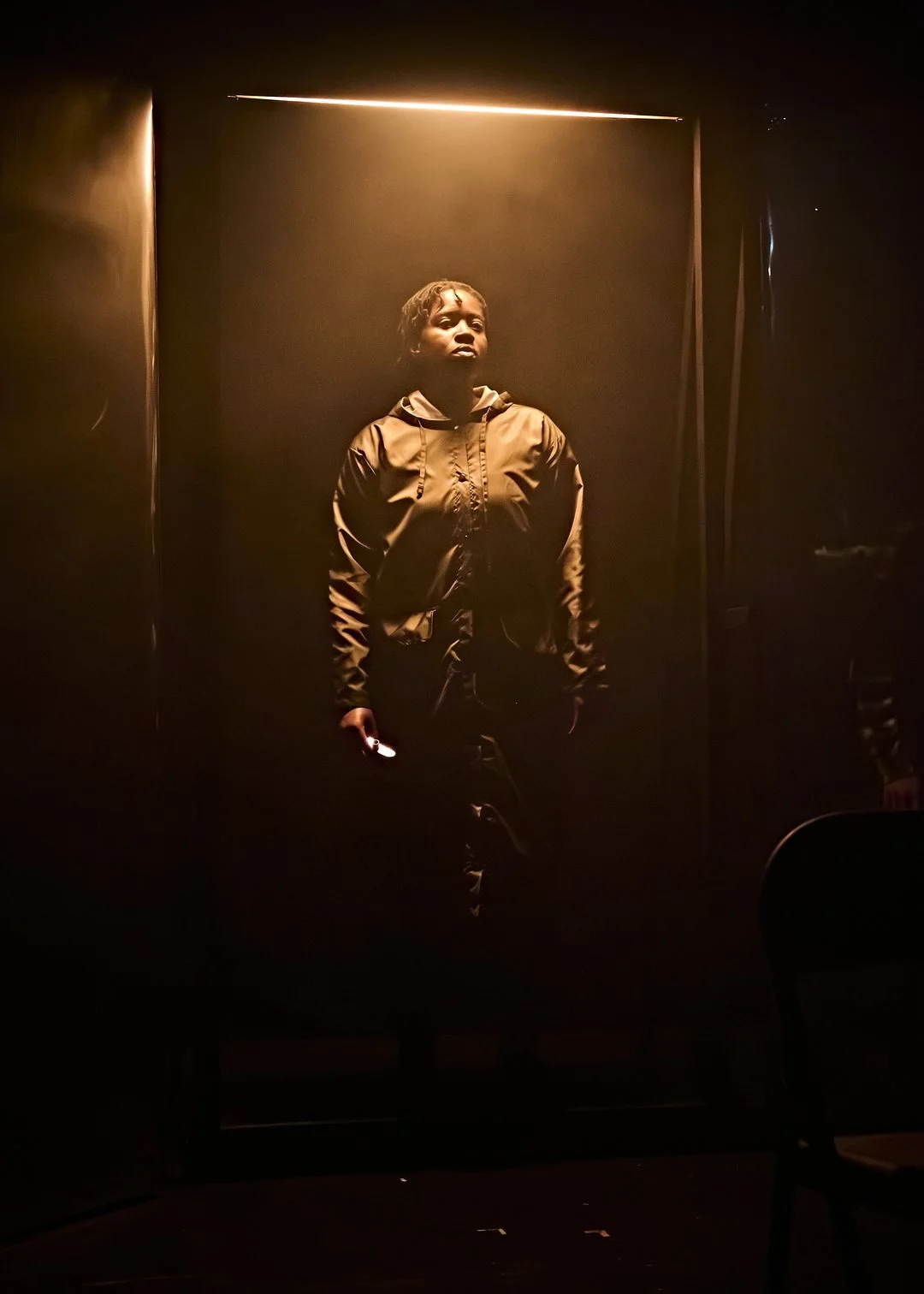 A woman standing in a dimly lit room with a wall behind her, dramatically illuminated by a strip of light from above.