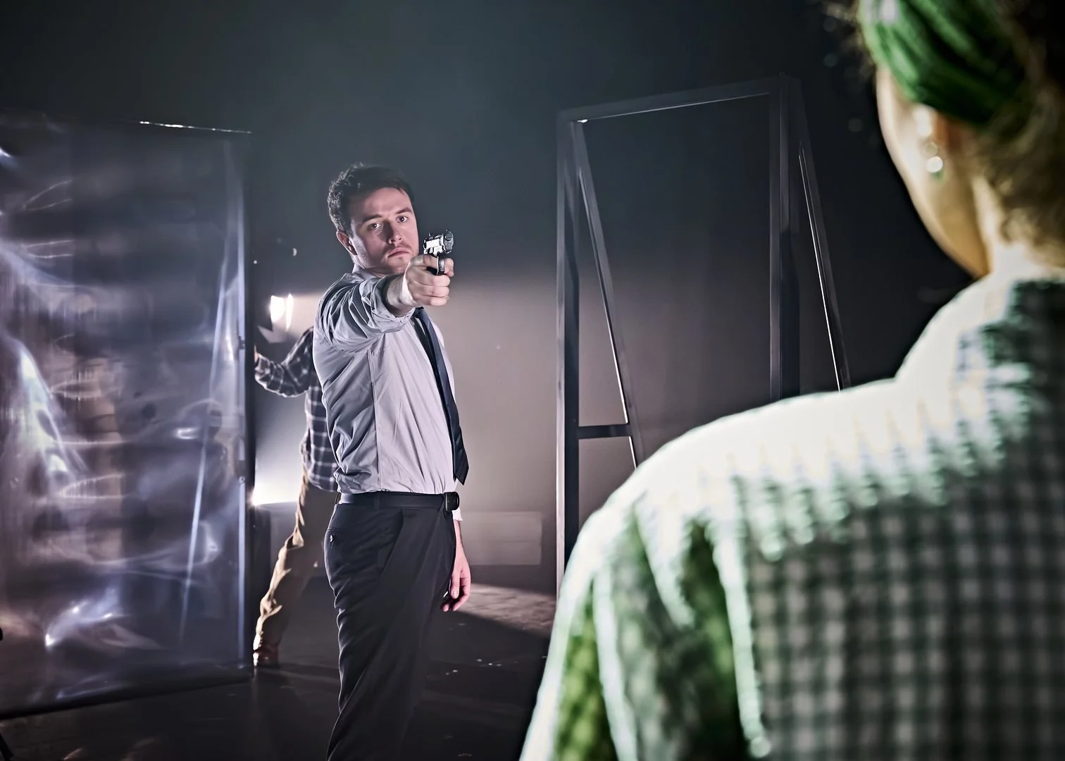 A man with dark hair in a suit and tie, holding a gun and aiming it at someone off-camera, in a tense scene with several people standing behind him in a dimly lit room.