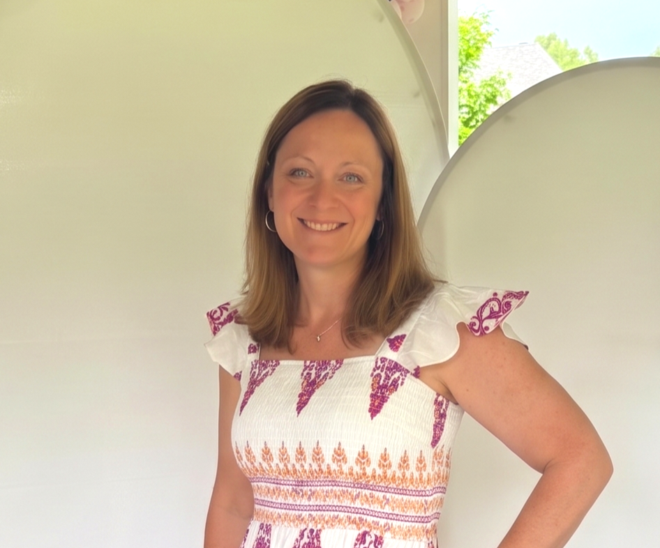 Woman standing in front of a white wall and window, smiling, wearing a white and purple patterned dress with ruffled sleeves.