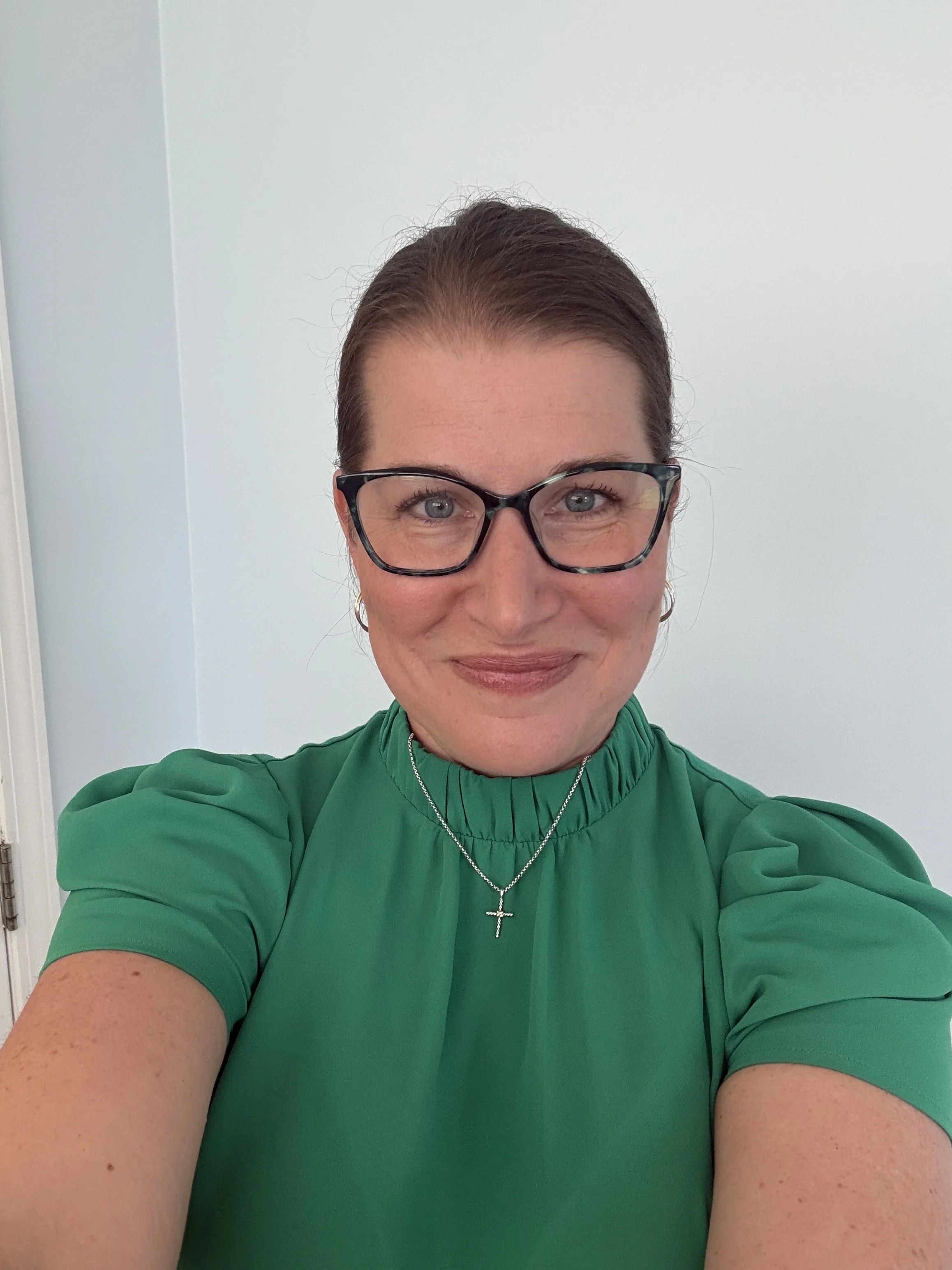 A woman with short brown hair, glasses, and a green top taking a selfie against a white wall.
