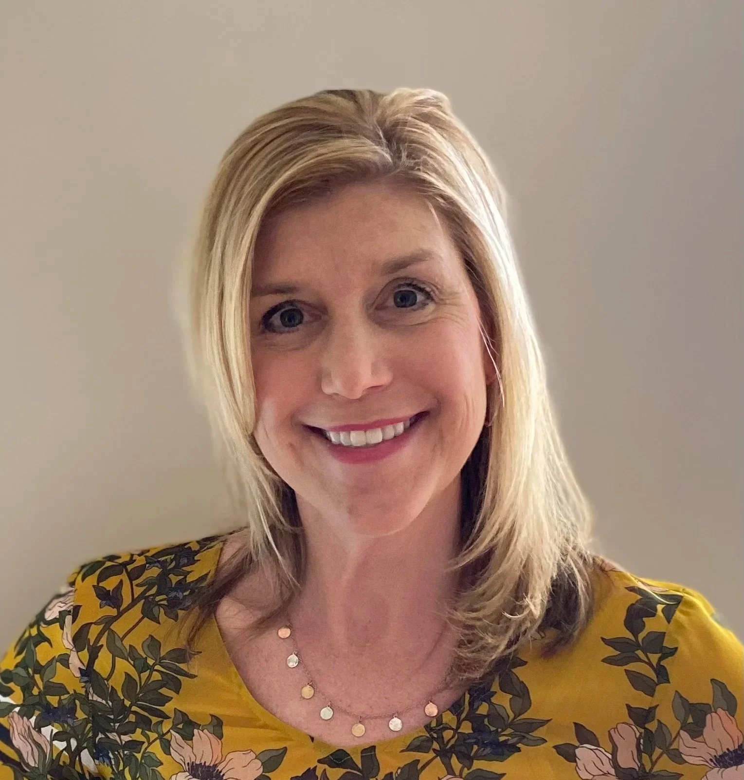 A woman with blonde hair, blue eyes, and a bright smile, wearing a yellow floral top and a layered necklace, posing against a plain, light-colored background.