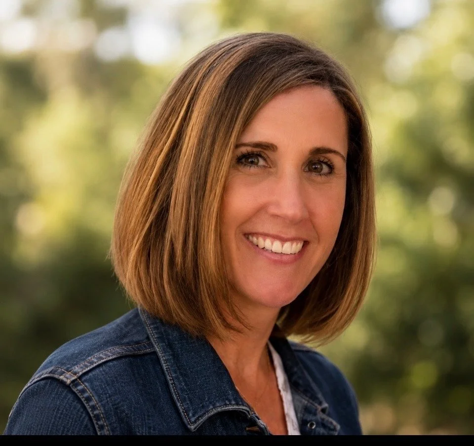 A woman with shoulder-length brown hair and a big smile, wearing a denim jacket standing outdoors in a natural setting with blurred green foliage in the background.