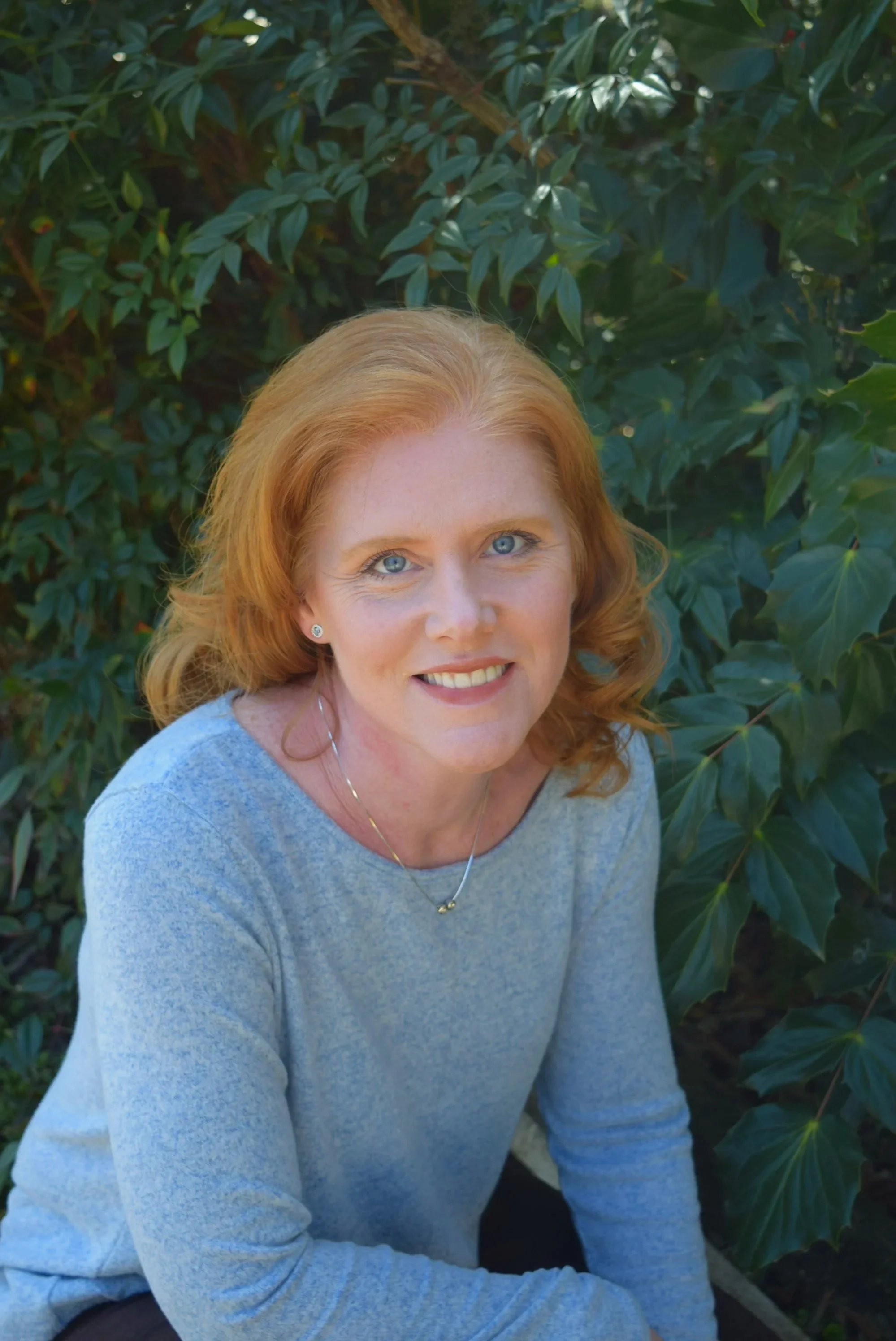 A woman with red hair and blue eyes smiling while sitting outdoors in front of green leafy bushes and trees.