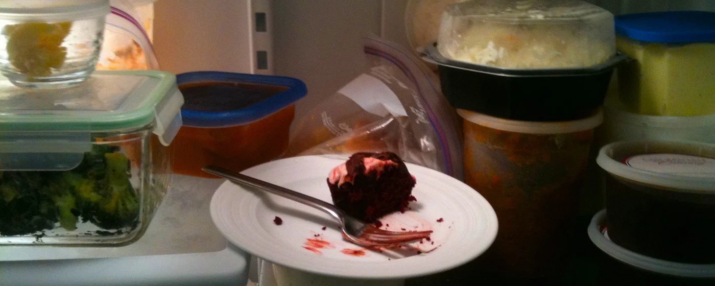 A close up photo of food in a refrigerator, mostly in containers. In the middle is a plate with a fork and a half eaten piece of chocolate cake with pink icing.