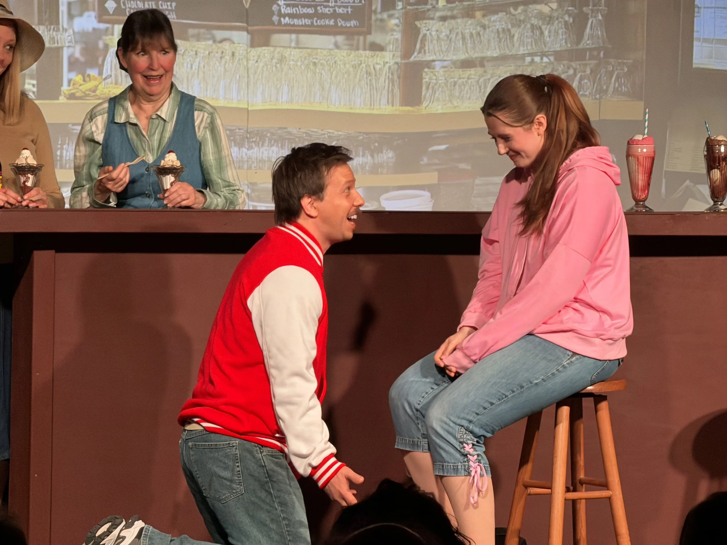 Two people kneeling and talking to each other in front of a half wall, with three people in the background standing behind the wall in a cafe or restaurant setting.