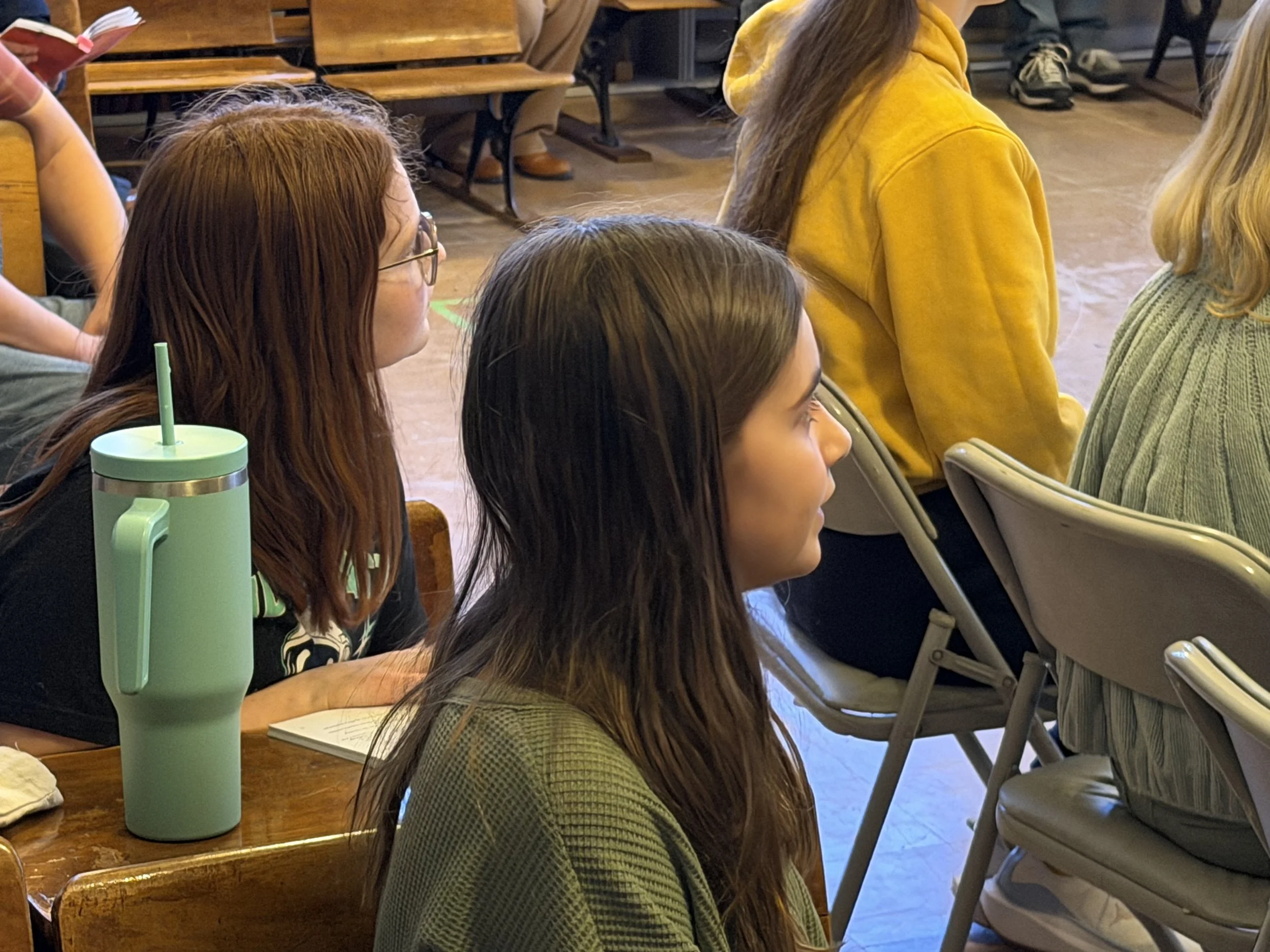 Multiple people sitting at a wooden table, some with notebooks, and a green water bottle with a straw in the foreground.