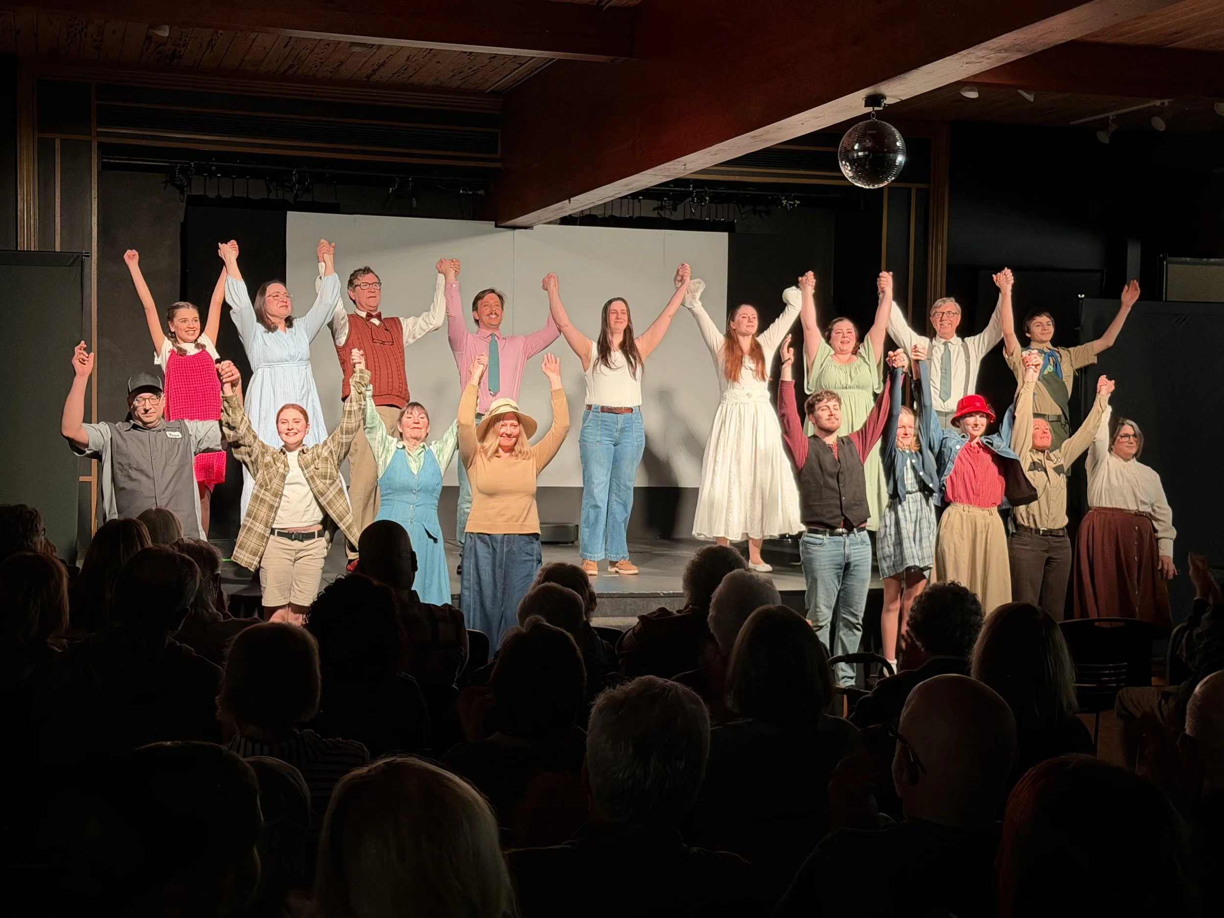 The image shows a group of performers on stage at the end of a theatrical production, holding hands and raising their arms in a gesture of victory or celebration. The audience is visible in the foreground, seated and watching the scene.