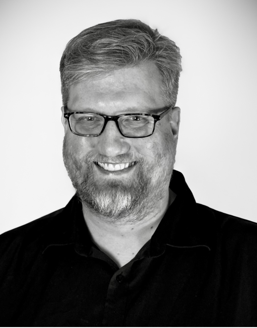 A black-and-white photo of a smiling man with glasses, sporting a beard and short hair, wearing a dark collared shirt.