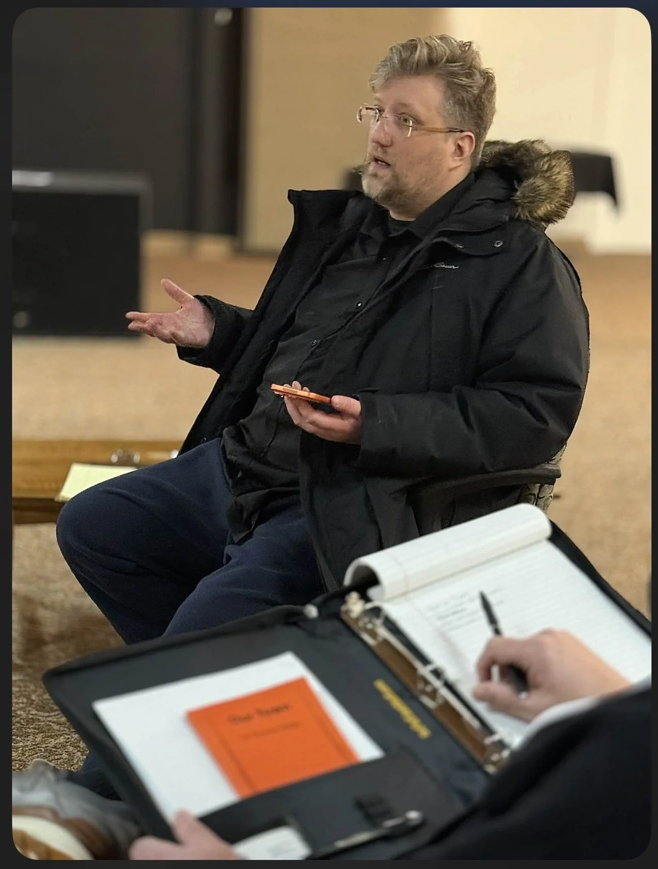 A man with glasses and a beard sitting and speaking in a discussion, wearing a black jacket with a fur-lined hood, in an indoor setting with a notebook and pen in the foreground.