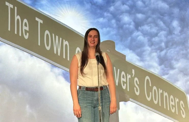 Woman with long brown hair standing in front of a microphone outdoors, with a large street sign and a partly cloudy sky in the background.