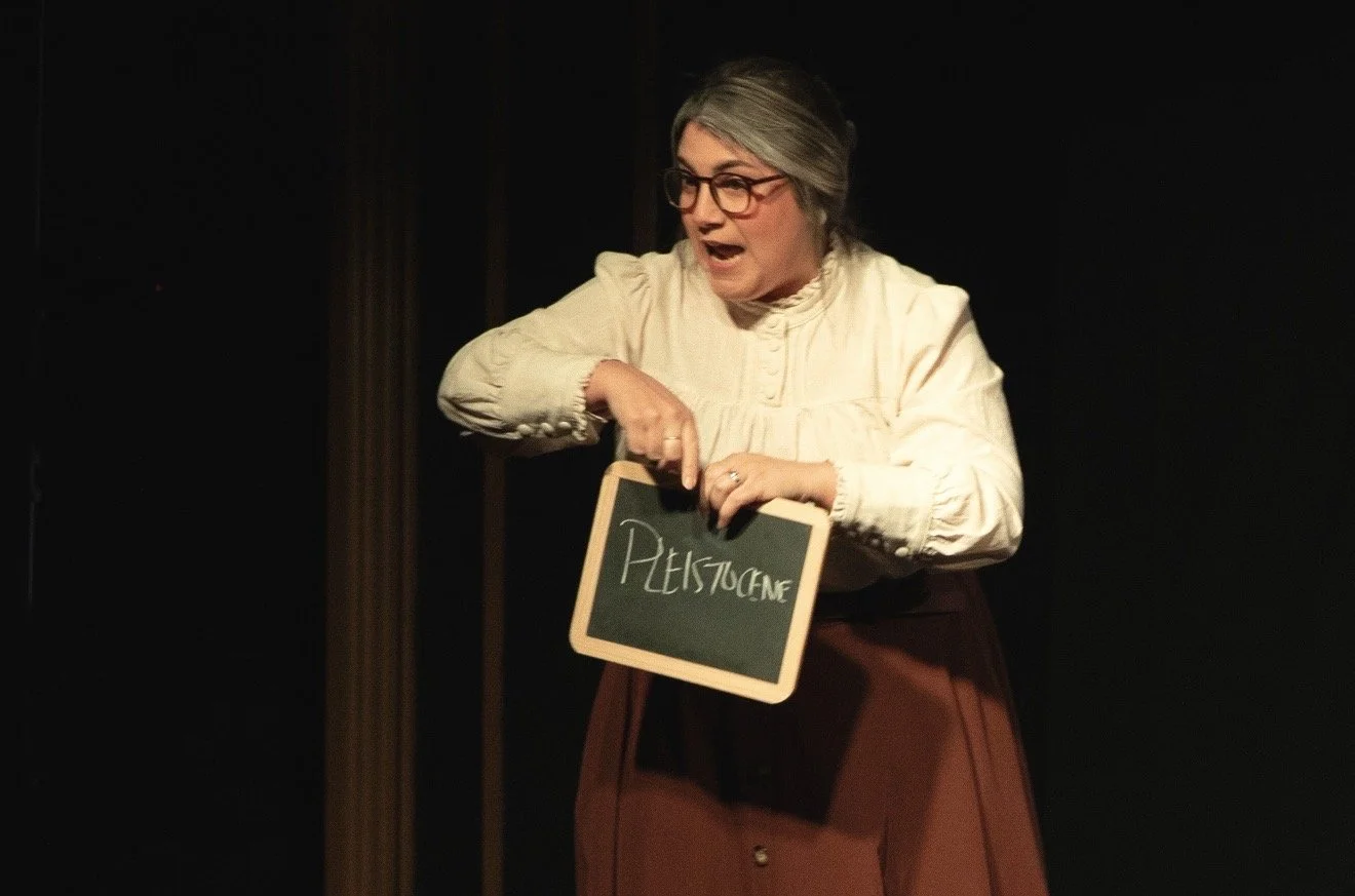 Woman performing on stage holding a small blackboard with the word 'PLEISTOCENE' written on it, wearing glasses and a cream-colored blouse.