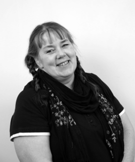 A black-and-white photo of a woman with light-colored hair in braids, smiling, wearing a dark scarf and a dark top with light trim.