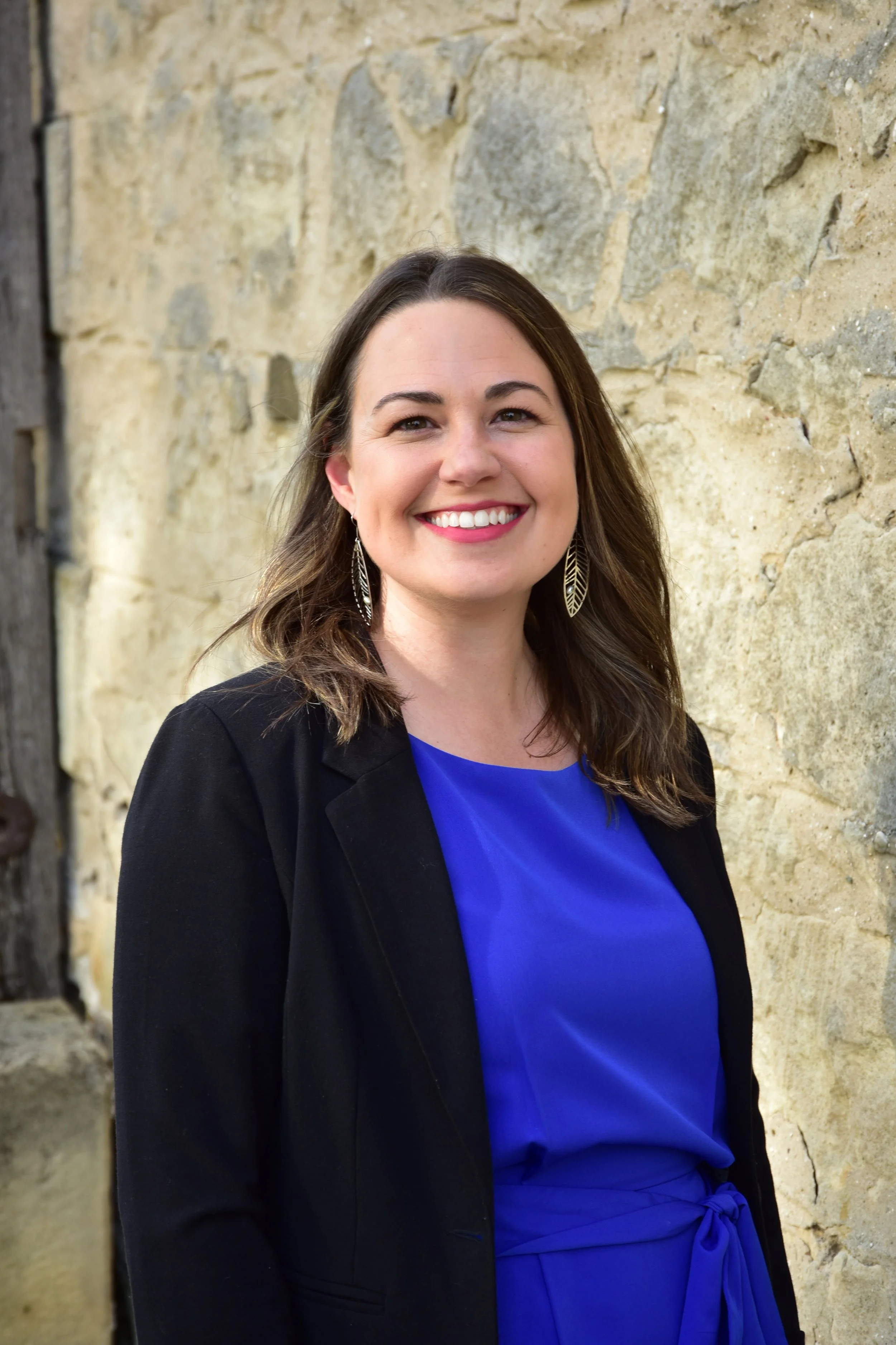 A woman with long dark hair, wearing earrings, a royal blue dress, and a black blazer, smiling and standing against a beige stone wall.