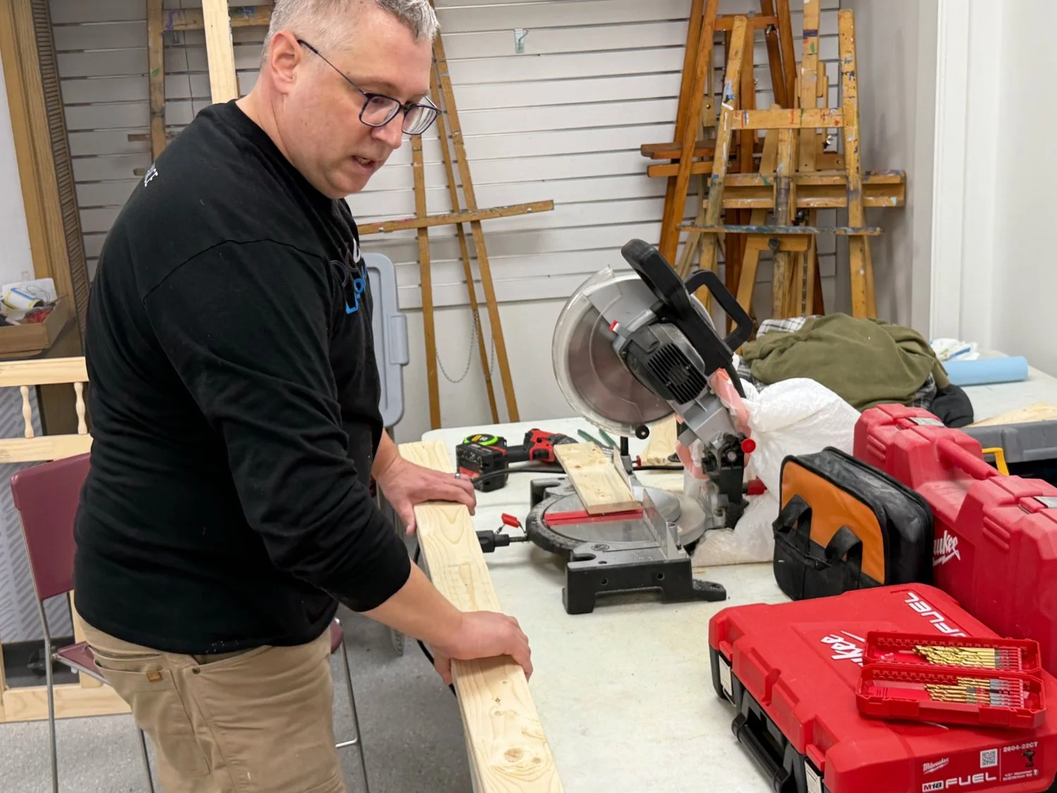 A man cutting a piece of wood on a miter saw in a workshop with various tools and ladders in the background.