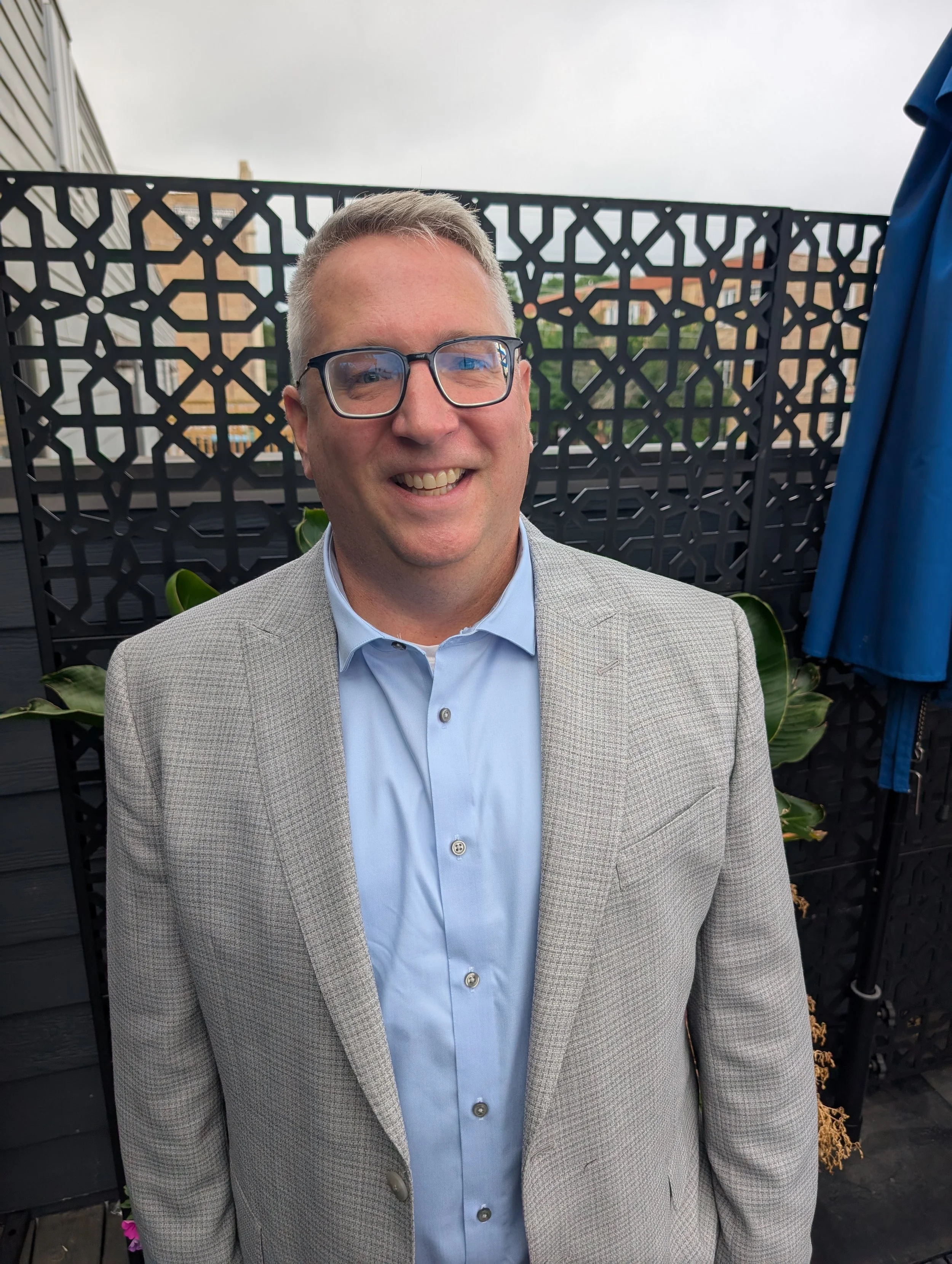 A man with short blonde hair, glasses, and a light-colored blazer smiling, standing outdoors on a balcony with a decorative black metal privacy screen and a cloudy sky in the background.