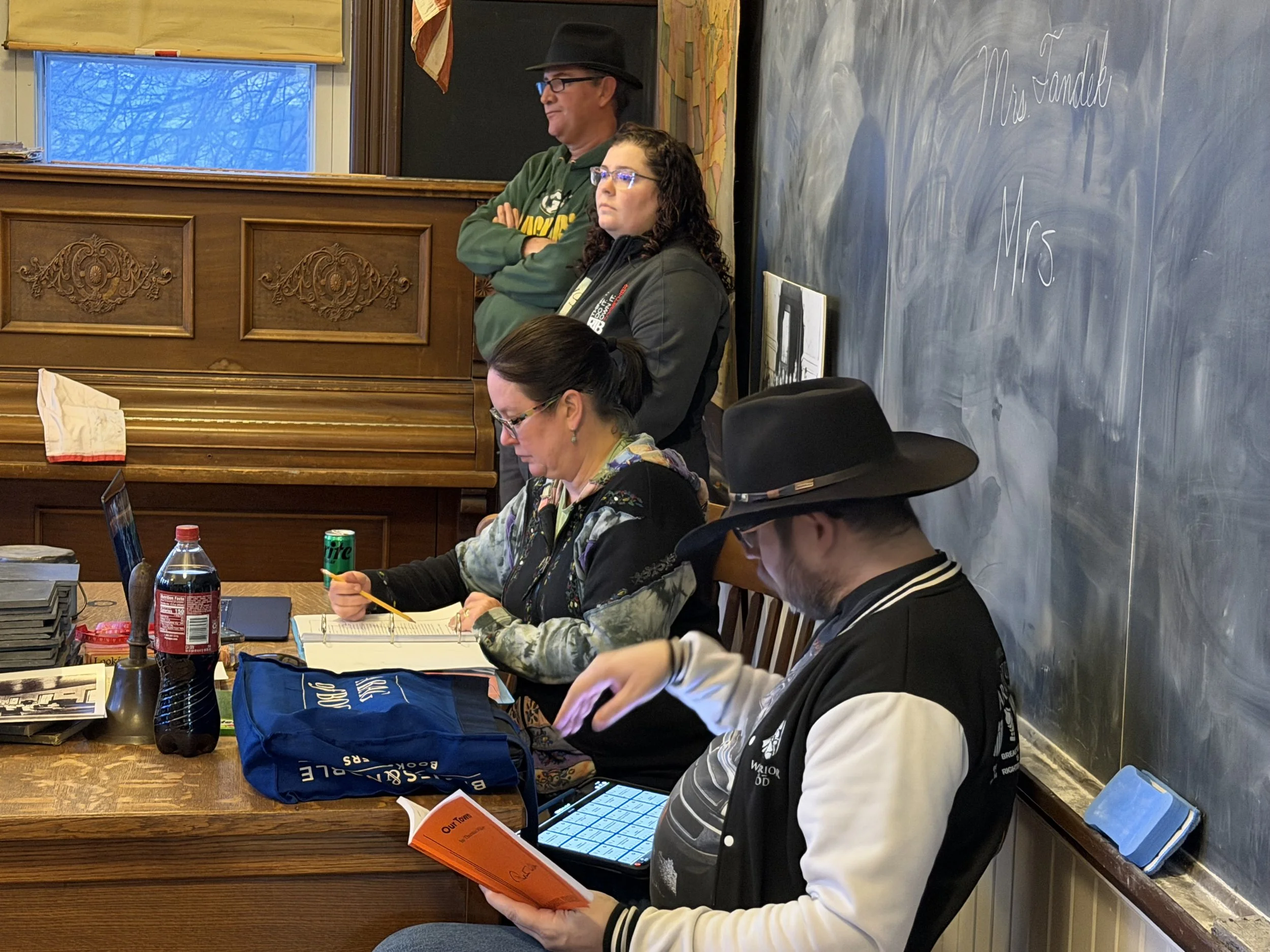 Group of four adults in a classroom, with three people sitting at a wooden table working on laptops and phones, and one standing at a chalkboard. The standing person is a woman with glasses, wearing a black jacket. One seated woman has glasses and is writing in a notebook, wearing a colorful tie-dye shirt. The person at the right is wearing a black and white varsity jacket and a black hat, reading a document and holding a tablet. The man standing is wearing glasses, a black hat, a green Wisconsin sweatshirt, and has his arms crossed. The classroom has a chalkboard with some writing, a window with ice and snow outside, and a table with various items including drinks and a blue bag.