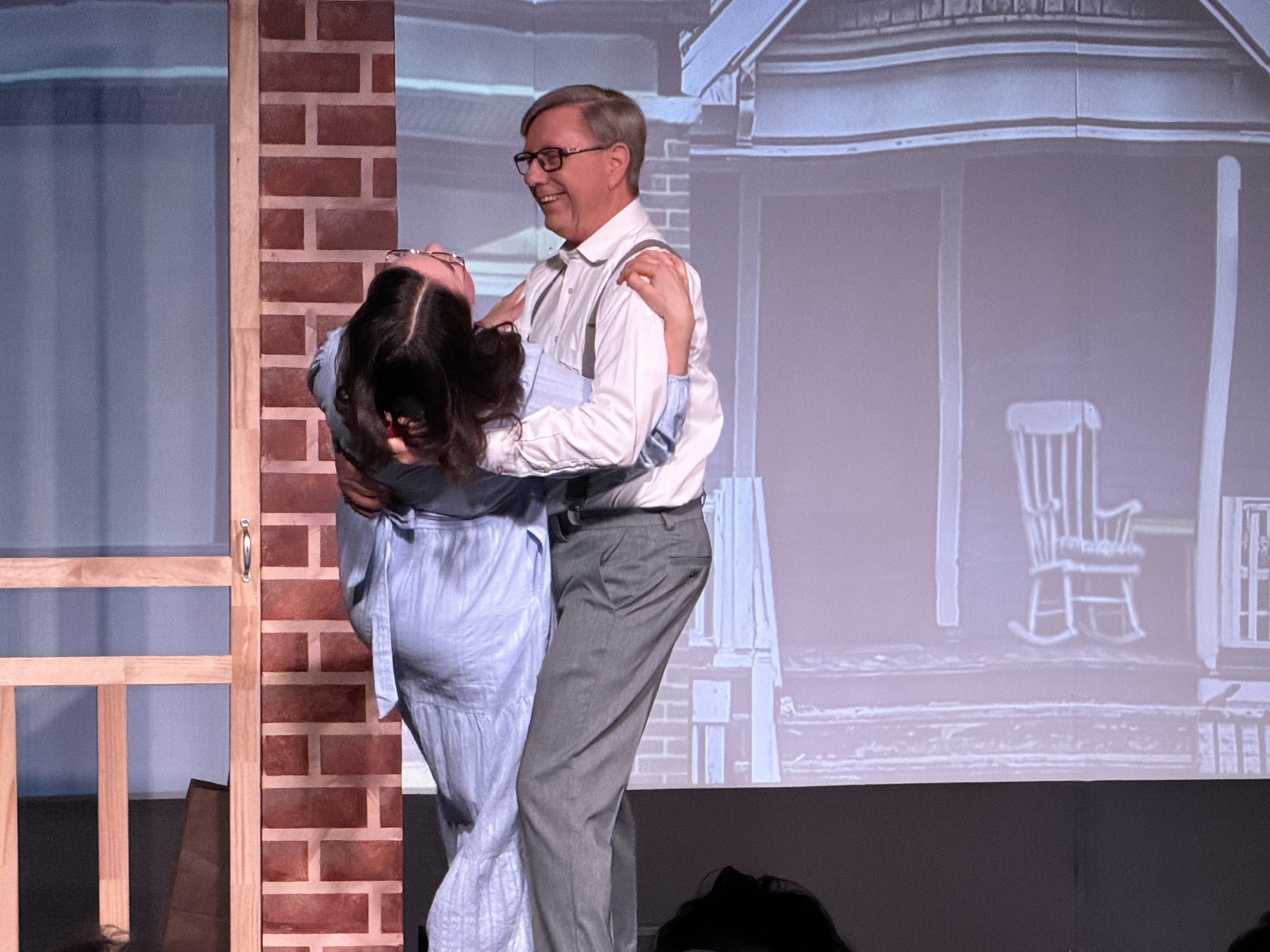 Two people, a man and a woman, sharing a joyful moment on stage, with the woman in an apron and the man in glasses and suspenders, standing in front of a background with a projected image of a porch or house exterior and a chalk outline of a chair.