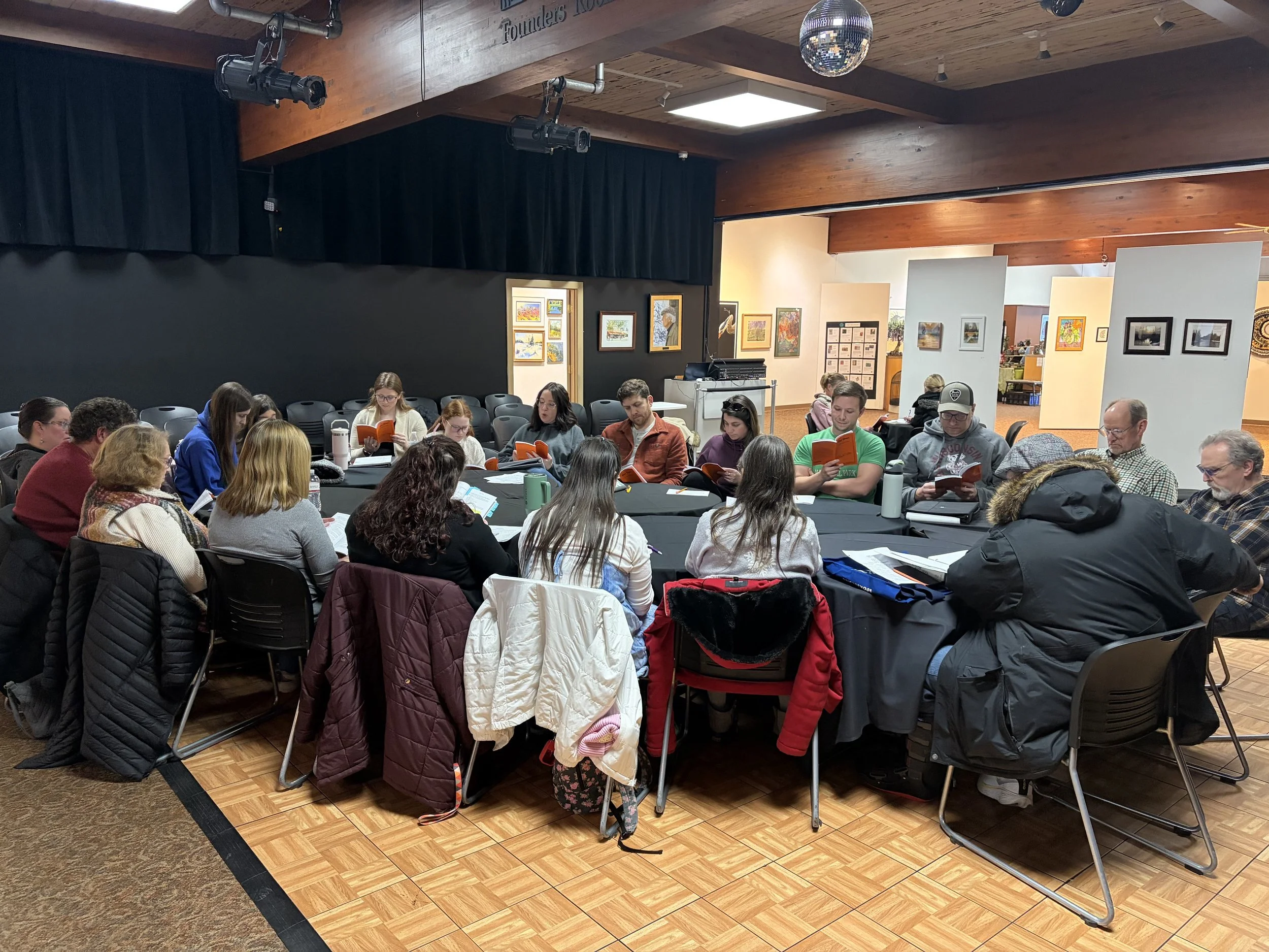 People sitting around a large table, reading books or documents, in an art gallery or exhibition space with artwork on the walls and a disco ball on the ceiling.