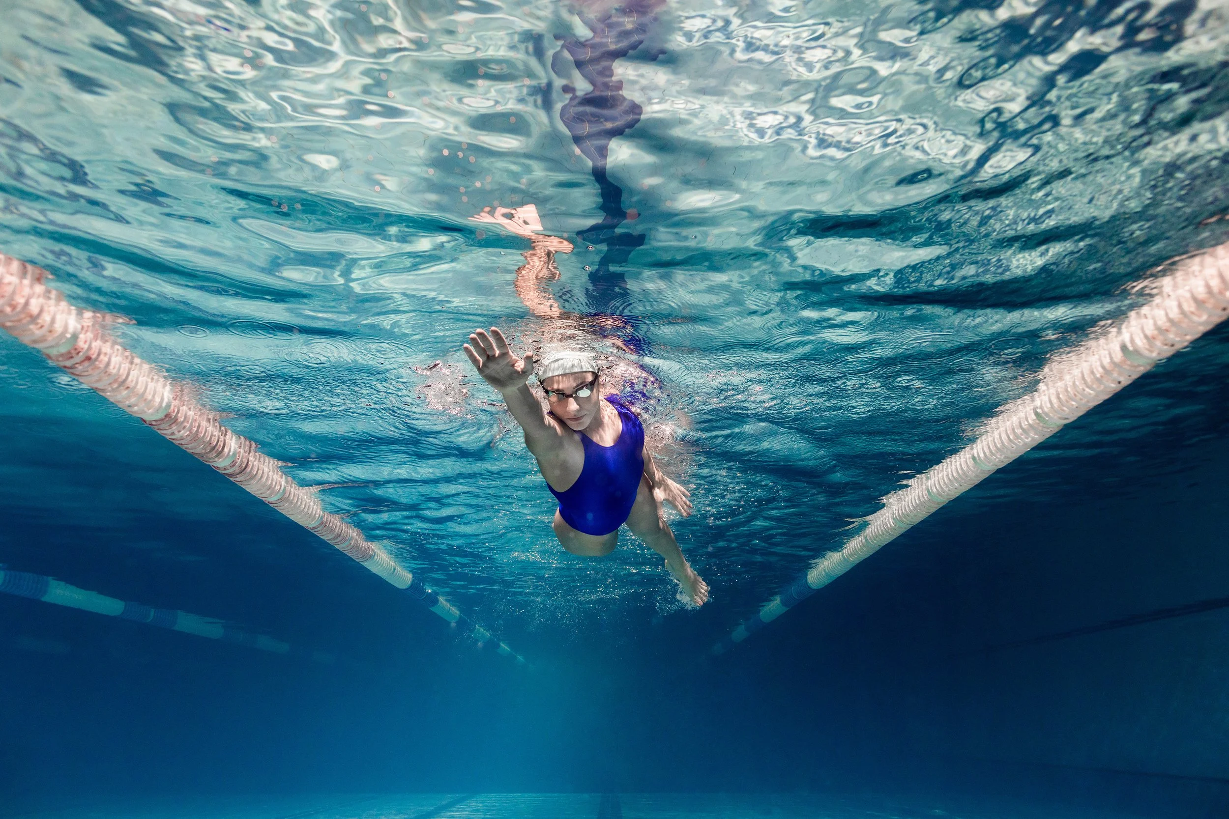 Sea Wisdom post-swim skincare brand image featuring a female swimmer in a blue swimsuit and goggles, underwater in a swimming pool, near the edge of the lane.