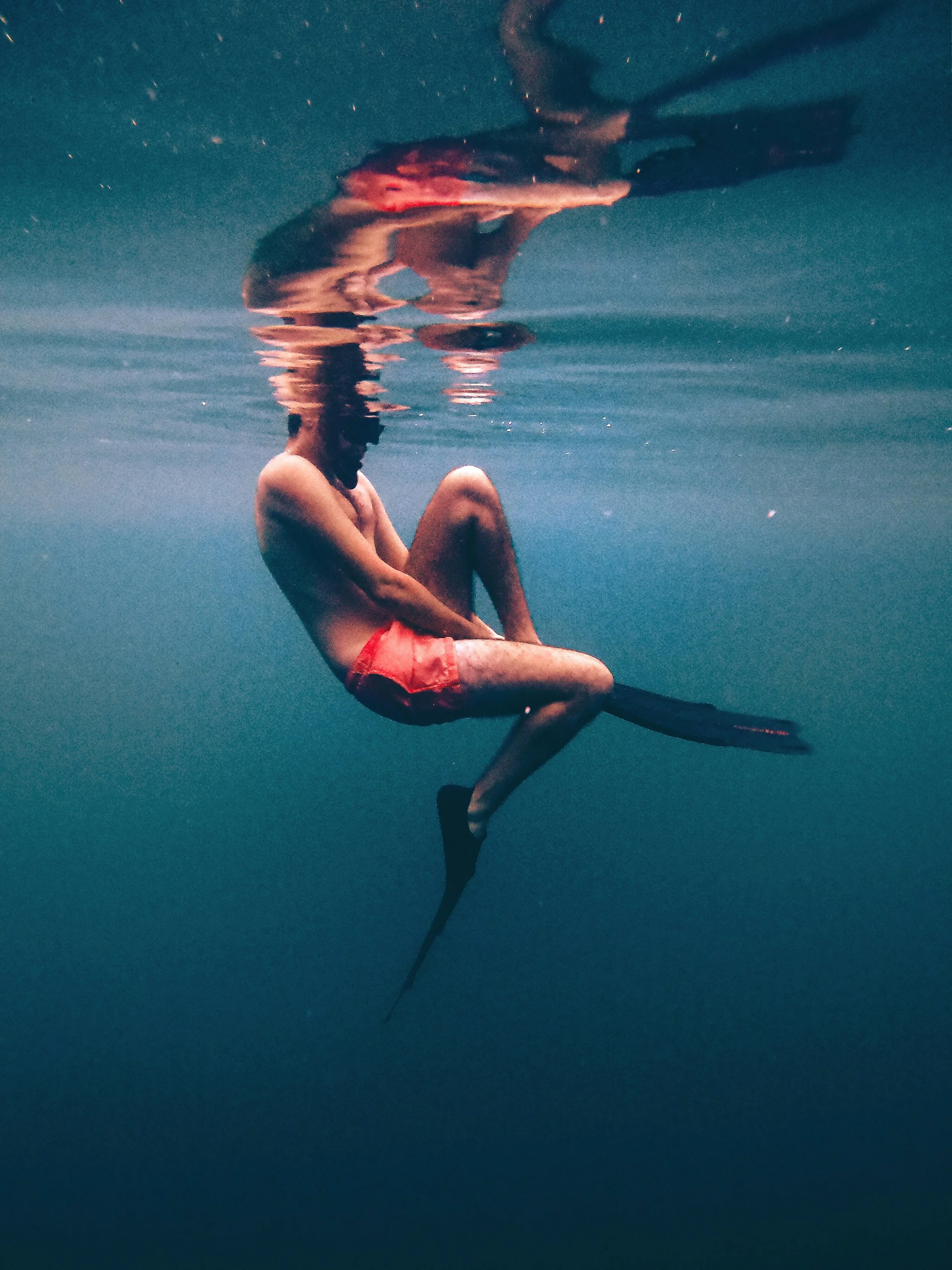 Sea Wisdom post-swim skincare brand image of a surfer sitting on a surfboard in the ocean, wearing red shorts, with their knees drawn up and their arms wrapped around their legs, looking down into the water.