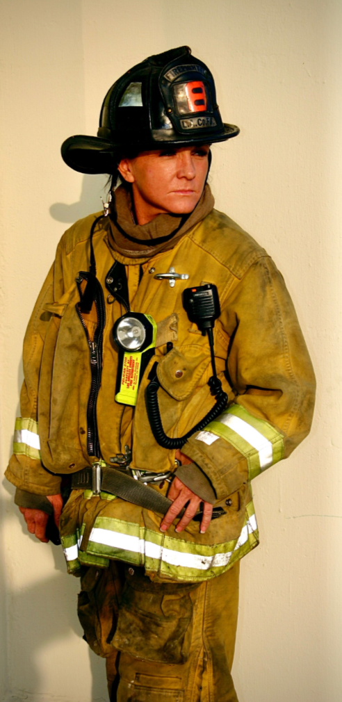 Firefighter in full gear, including helmet, jacket, and reflective cargo pants, standing against a plain wall.