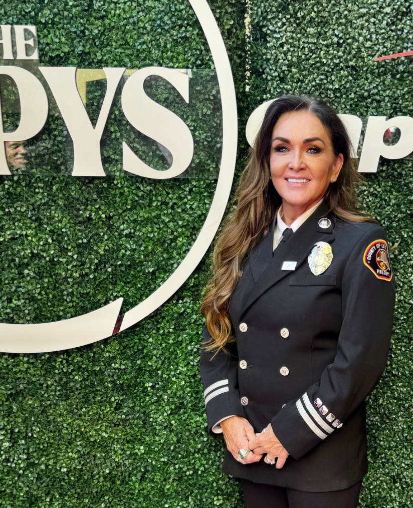 A woman in a firefighter uniform standing in front of a green hedge wall with a large sign that reads 'The Pysi'.