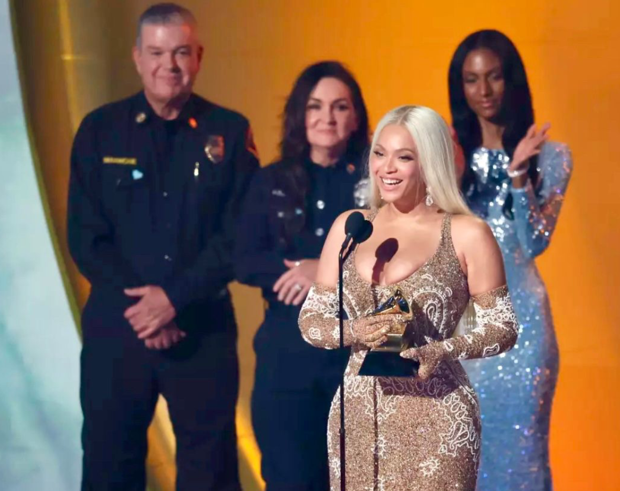 A group of four women and one man standing on stage, with one woman at the front holding a Grammy award. The woman in front is wearing a sparkly gold gown and smiling. Others are dressed in black police uniforms and a silver dress, standing behind her against a yellow background.