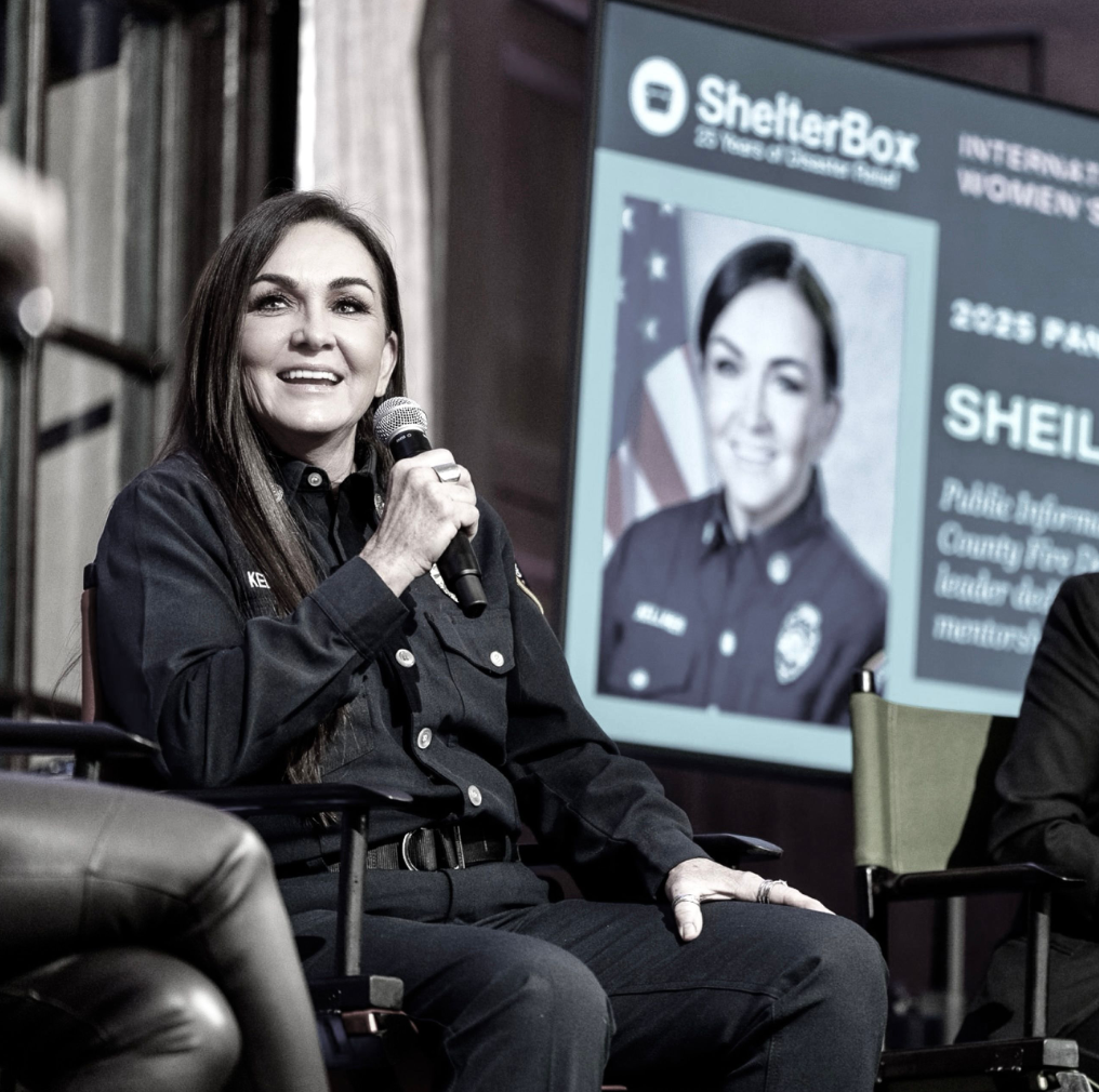 A woman police officer sitting on a chair, holding a microphone and speaking at an event, with a large screen in the background displaying her picture and event information.